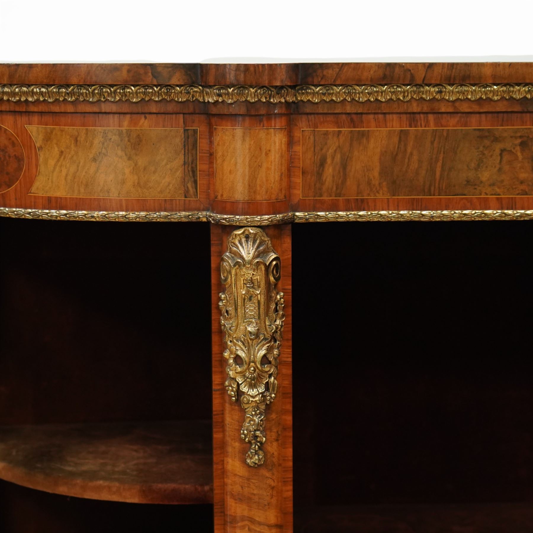 Late Victorian figured walnut credenza, the shaped demi-lune top with cross-banding and stringing over an inlaid frieze with foliate gilt-metal mounts, fitted with a central geometric-inlaid panelled door, flanked by convex-glazed side-cabinets with a gilt metal slip and outer-banding, the uprights surmounted by gilt-metal cartouche mounts with cascading foliage details, raised on a shaped skirted base with central female-mask gilt mount