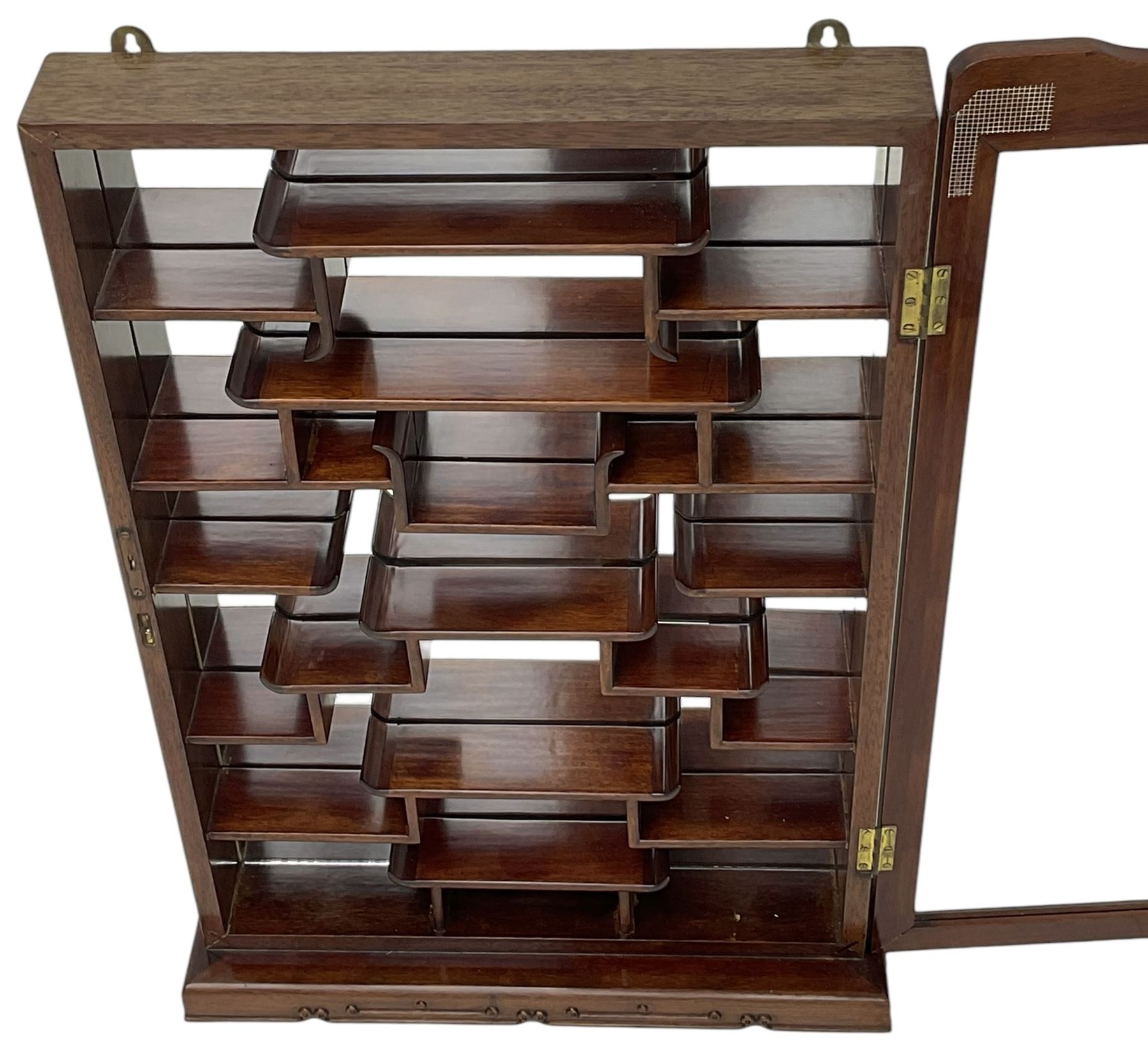 Chinese hardwood wall hanging curio cabinet, the mirror interior fitted with stepped shelves, enclosed by single glazed doors, decorated with carved scrolling foliage 