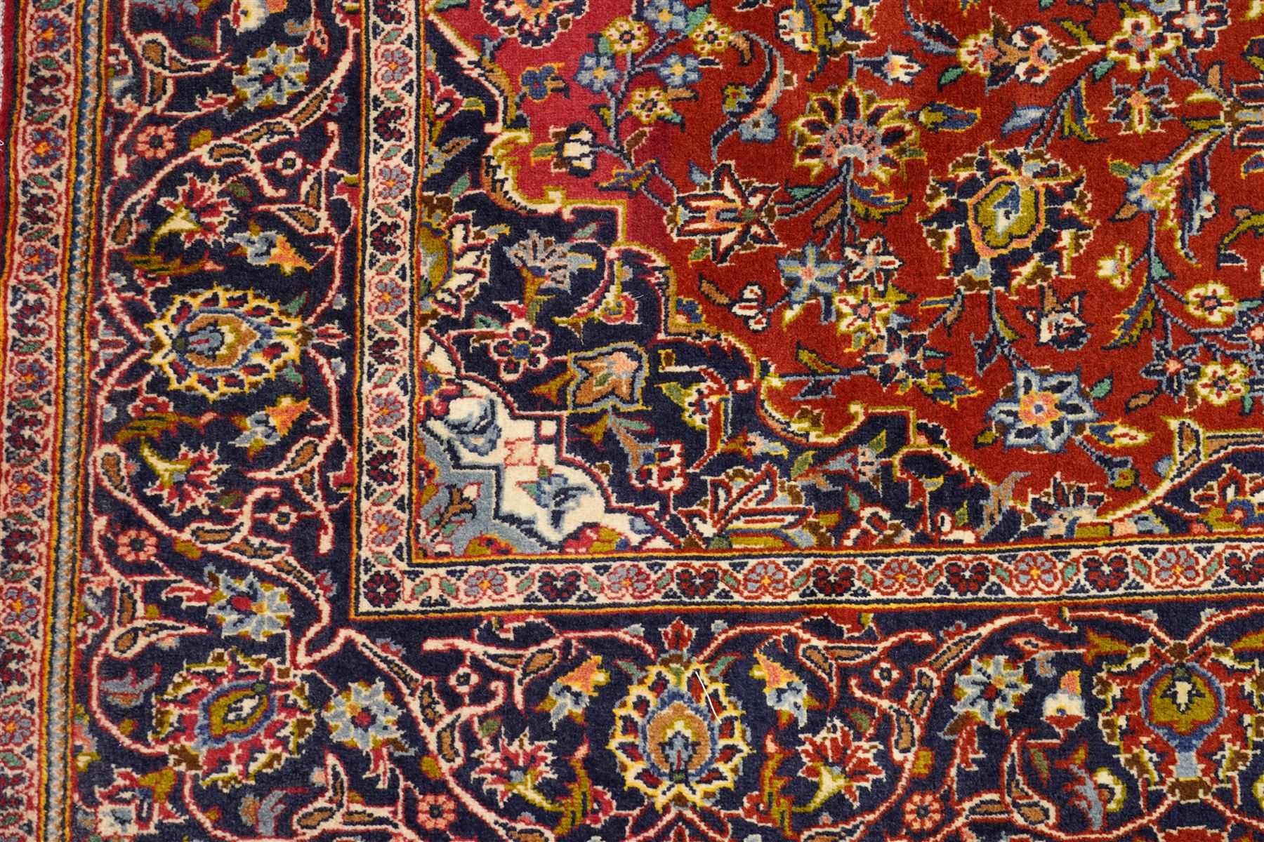 Central Persian Kashan hand-knotted rug, deep red field with central cusped ivory medallion with an all-over arrangement of scrolling floral vines, palmettes and flowering sprays within conforming spandrels, principal navy border with a continuous meandering vine of palmettes and stylized floral motifs, enclosed by multiple narrow guard borders, with cotton fringe ends 