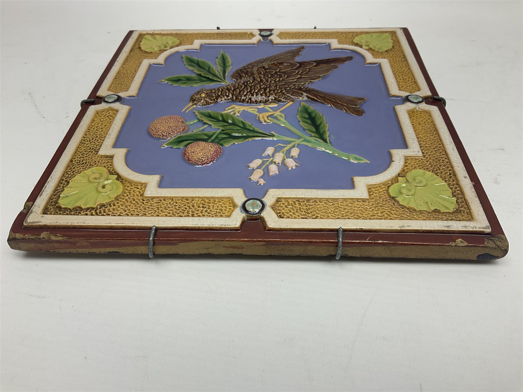 19th century Minton & Co majolica tile, decorated in relief with a bird upon a fruiting branch against a blue ground, with textured ochre border and anthemion to each corner, impressed mark verso, H19.7cm
