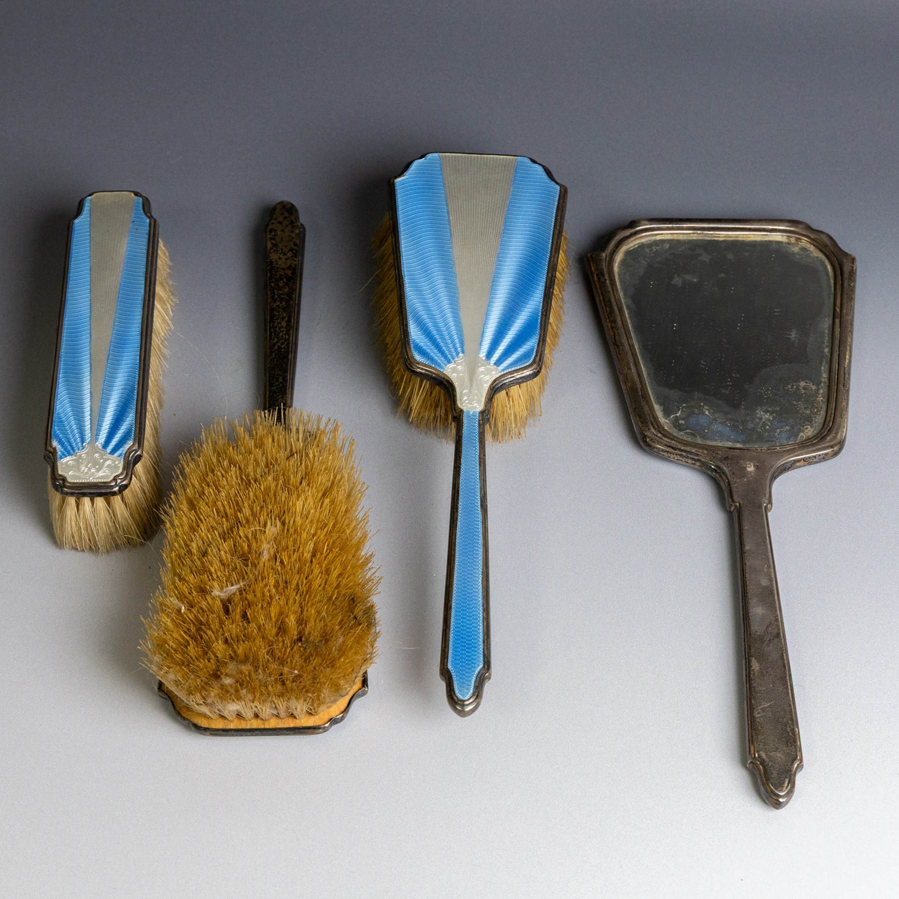 Art Deco four piece dressing table set, comprising hand mirror, two hair brushes and clothes brush, each with blue and white guilloche enamel decoration, hallmarks worn and indistinct