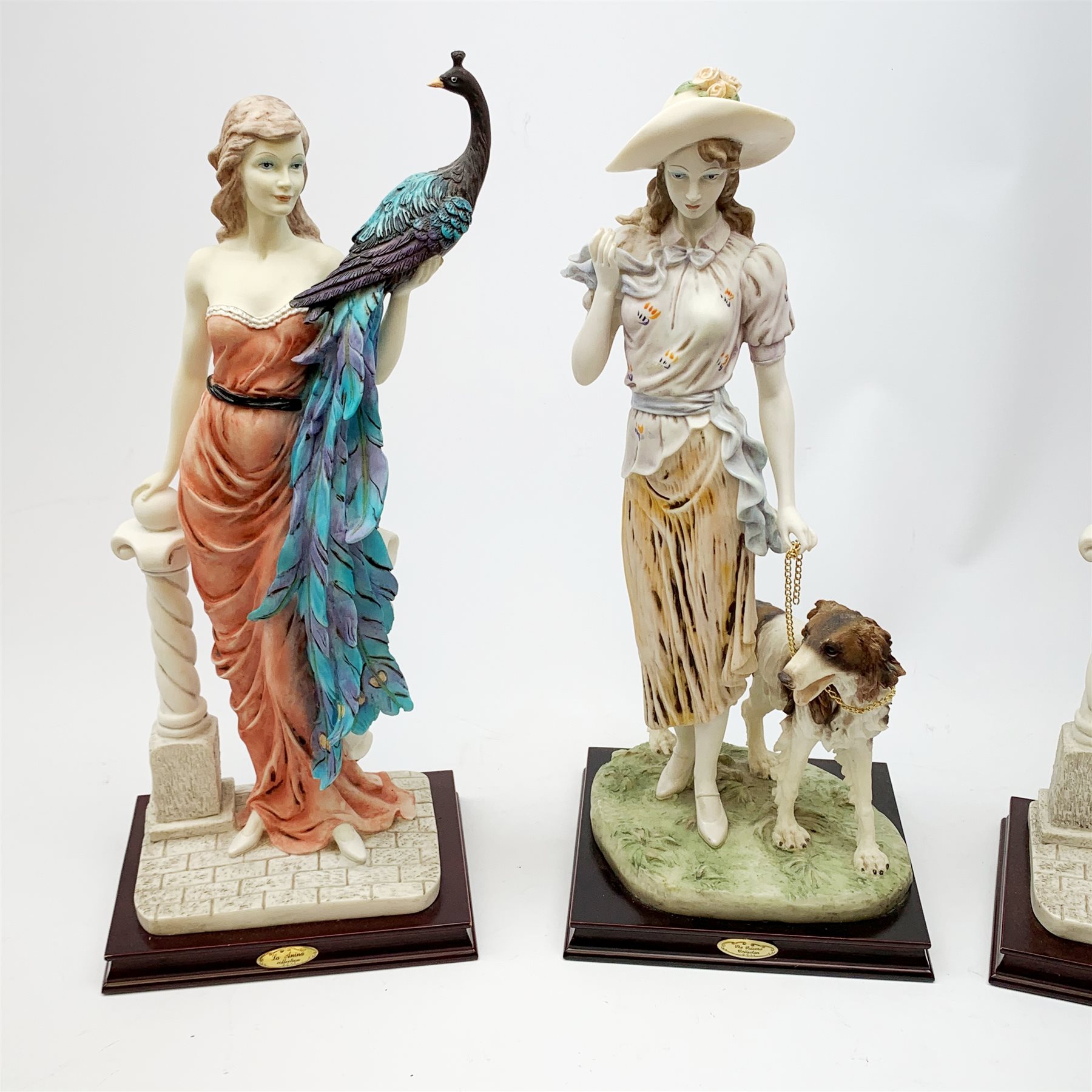  A pair of La Anina Collection figurines, each modelled as a female figure in pink dress supporting a peacock on one arm, and resting the other upon a classical column, H51.5cm, together with another similar figurine modelled as a female figure with hound at her side.   