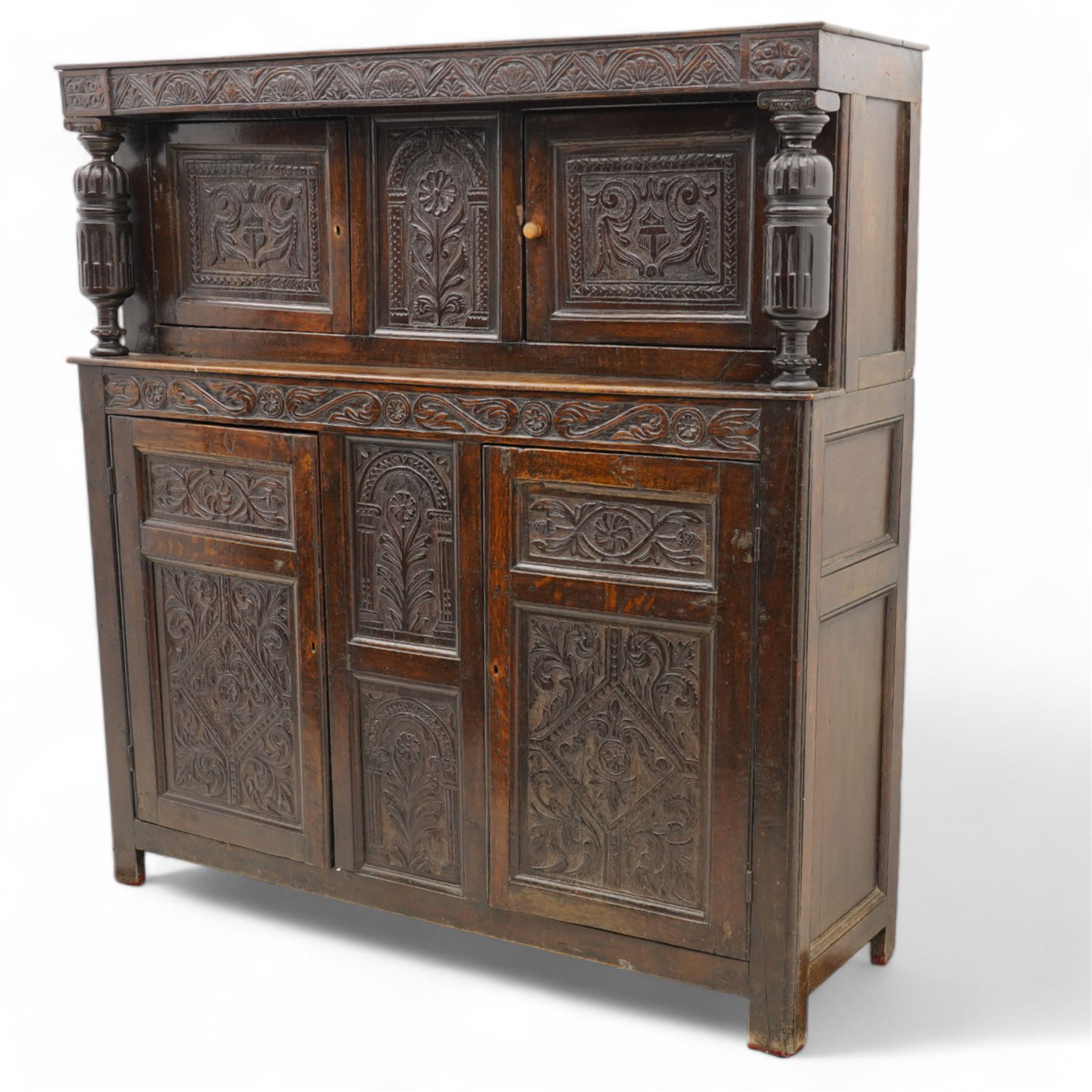 William & Mary design oak court cupboard, lunette carved frieze rail on turned and stop-fluted columns, the upper section fitted with two panelled doors carved with scrolling foliage and shield within a chip-carved border, central panel carved with arcade surrounding flower, foliate S-scroll lower frieze rail over two further cupboards and central panels, each carved with lozenges and curling foliage, moulded and pegged frame, terminating to stile supports 