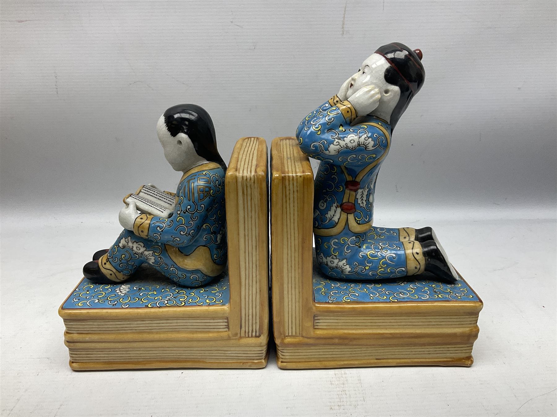 Pair of 20th century Chinese figural bookends, modelled as children in traditional blue dress sat reading and kneeling upon stacks of books, H22cm