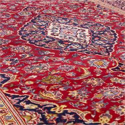 Central Persian Kashan hand-knotted rug, crimson red field with central ivory and indigo floral medallion surrounded by an all-over arrangement of finely drawn palmettes, flowering vines, and rosettes, with corresponding navy blue spandrels enclosed by a floral main border with multiple guard stripes, with cotton fringe ends    