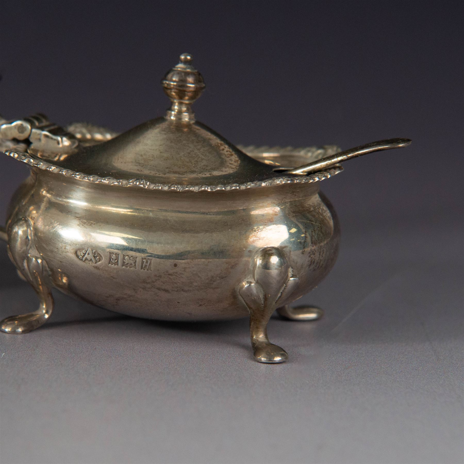 Mid 20th century silver three piece cruet set, comprising pepper shaker, open salt, mustard pot with cover and two condiment spoons, all of oval form with oblique gadrooned rims and upon four paw feet, hallmarked Adie Brothers Ltd, Birmingham 1957, with blue glass liners