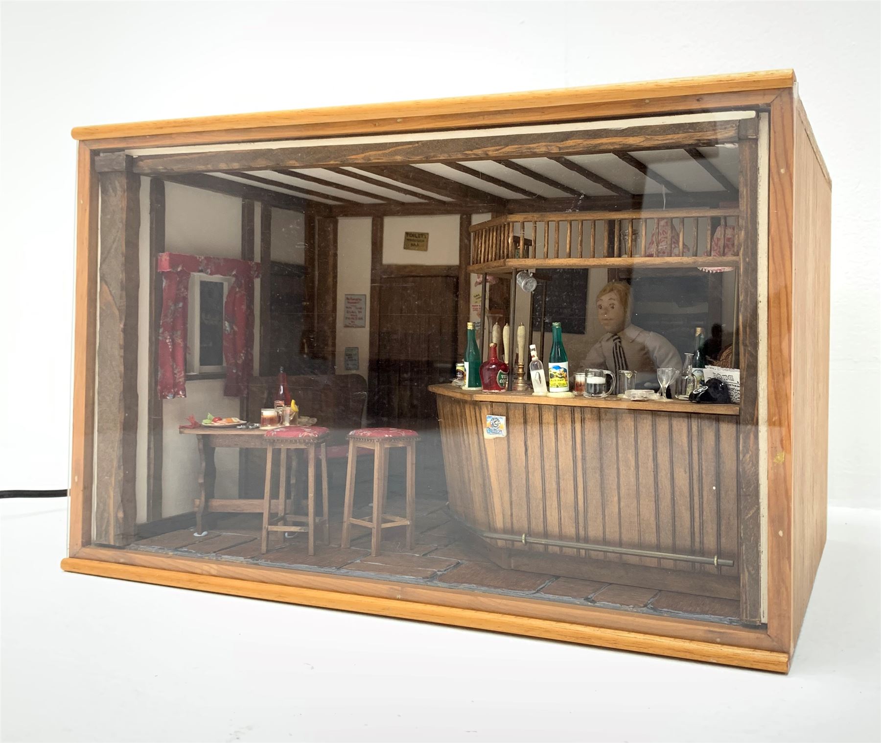 Dolls house scale room setting of a public house bar with tiled floor and beam and plaster ceiling and walls, bench seat, stools and table set out with a meal and drinks, large planked finish canted bar well stocked with food, drinks, bottles, glasses etc and bartender figure behind; electrically illuminated with sliding perspex access panel to front W38cm D28cm H24cm
