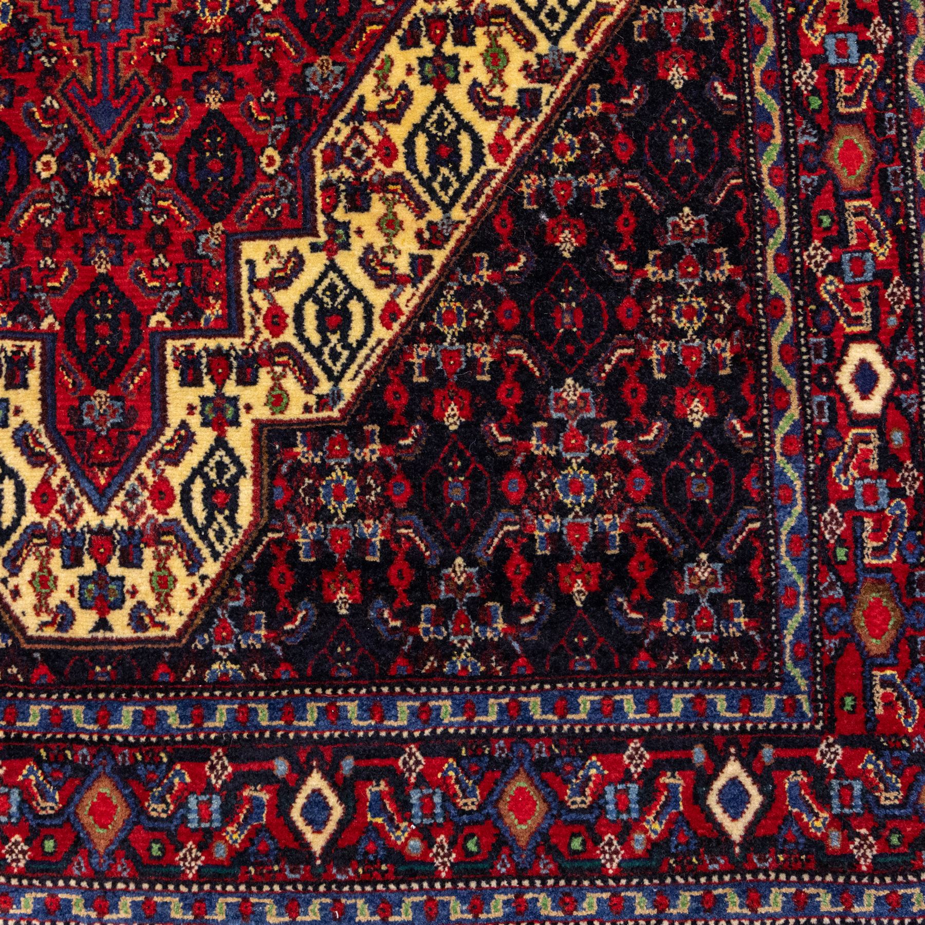 North West Persian Senneh hand-knotted rug, dark indigo field with a central bold stepped lozenge medallion of madder red and deep blue, enclosed within an ivory serrated contour forming a cruciform outline, medallion with repeating boteh and floral devices, field decorated with an all-over Herati pattern of rosettes enclosed by angular leaves principal border of madder red ground decorated with alternating floral heads, flanked by narrow guard stripes, with cotton fringe ends 