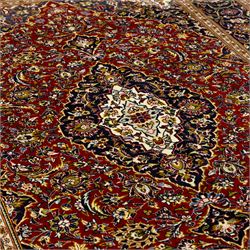Central Persian Kashan hand-knotted carpet, crimson ground, with a central ivory shaped floral medallion with pendants, surrounded by dense scrolling foliate and floral motifs incorporating rosettes, palmettes and delicate vine work, enclosed by ivory spandrels and a dark navy border with repeating palmettes and meandering vines, flanked by floral guard stripes, with cotton fringes
