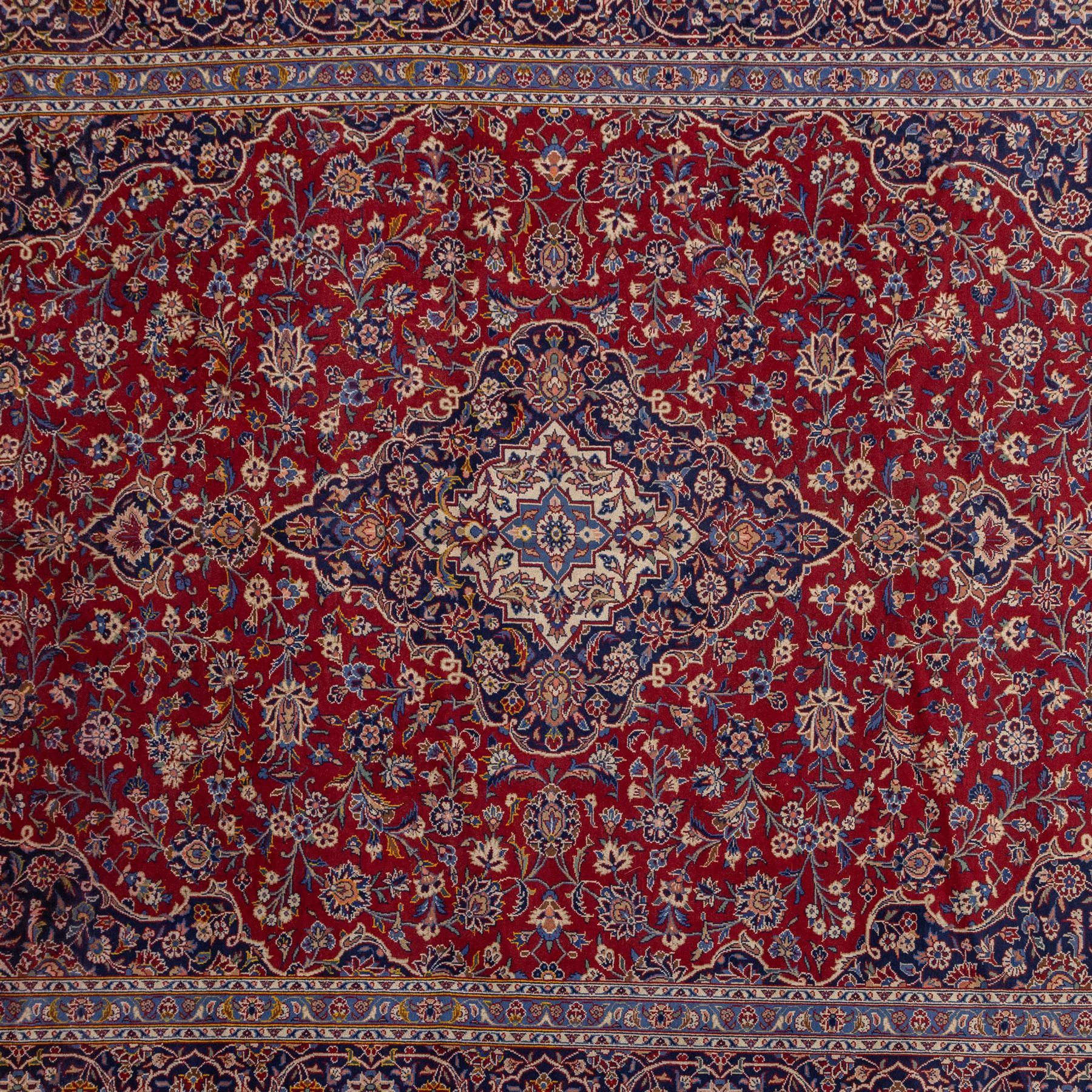 Central Persian Kashan hand-knotted carpet, madder red field with central cusped medallion in navy and ivory enclosing a floral rosette and radiating scrolling arabesques and palmettes, across a densely ornamented ground of finely drawn vinery enriched with a profusion of flowering sprays and stylized blossoms, spandrels in deep indigo and principal border of navy ground decorated with a continuous palmette and vine-scroll flanked by multiple guard stripes, with cotton fringe ends