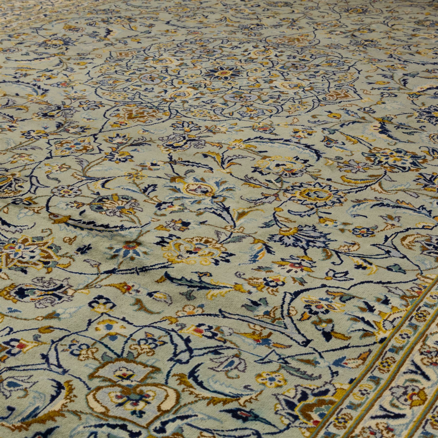 Central Persian Kashan hand-knotted carpet, pale celadon blue ground, decorated with an all-over central floral and foliate design with rosette medallion incorporating scrolling vine work, palmettes, and stylized blossoms  highlighted in tones of indigo, ivory, gold, and powder blue, enclosed by a cream ground border, flanked by multiple guard stripes, with cotton fringe ends 