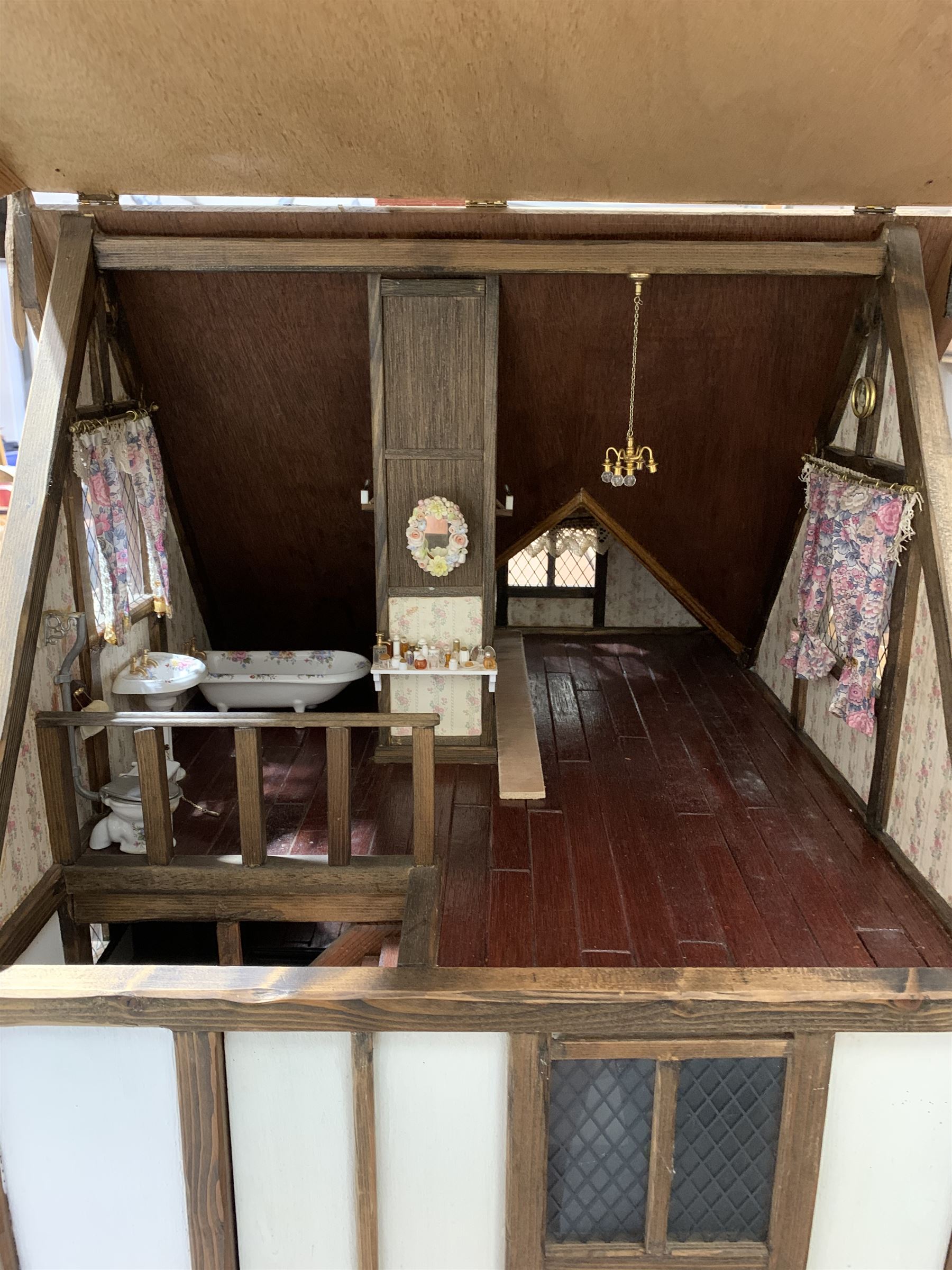 Gerry Welch for Manorcraft Tudor style wooden doll's house, of three-storey multi gabled form with mock beam and plaster walls and simulated leaded glass windows under a felt tiled roof, boarded floors and stairs to each room, curtains to most windows, front entrance door, rear door and single storey extension for kitchen. Furniture includes fitted floral bathroom suite to top floor, well stocked Welsh dresser with copperware etc, tapestry style wall hangings, knole settee, tin bath, metal range, fire-surround, wooden three-piece suite etc; on shaped cobbled base W91cm D61cm H91cm