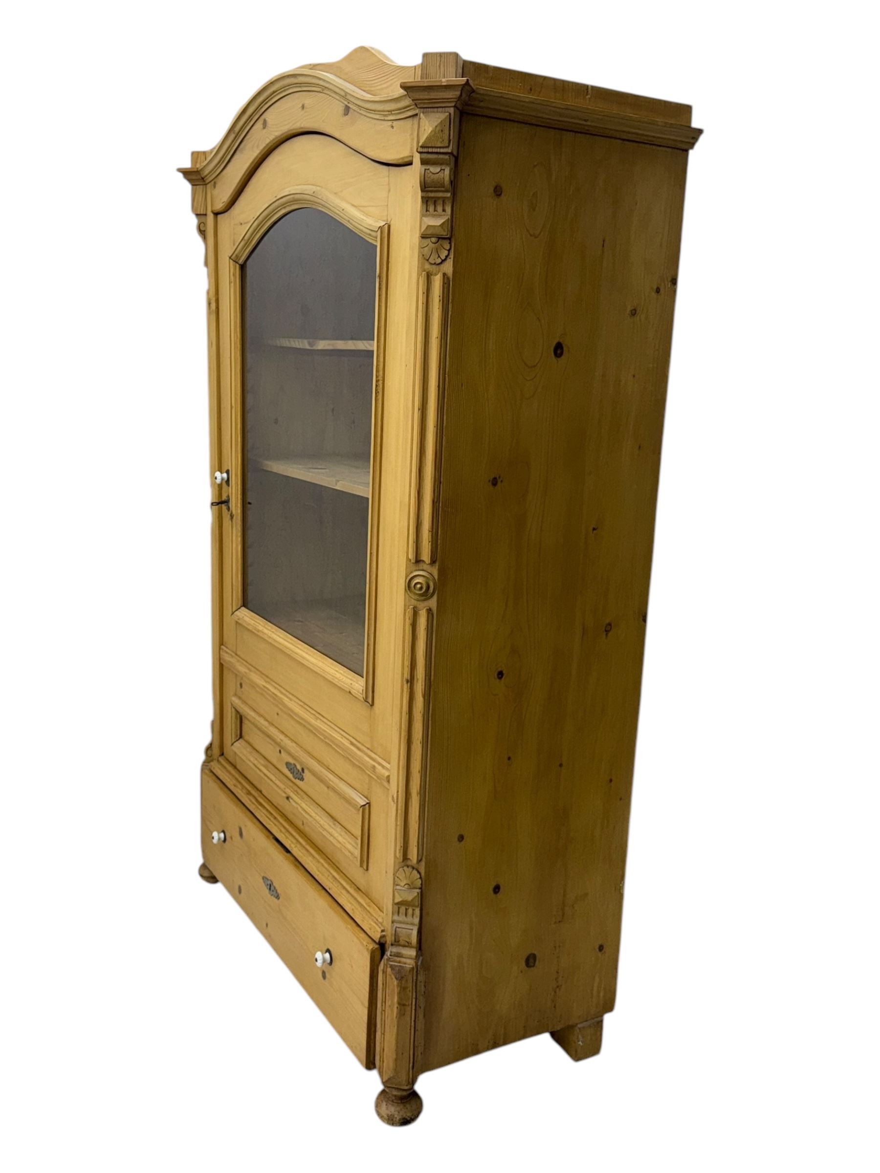 Late 19th century Continental pine vitrine, shaped and moulded arched cornice over a single glazed door enclosing adjustable shelves, flanked by carved pilasters with foliate and geometric motifs, fitted with single moulded drawer, raised on compressed bun feet