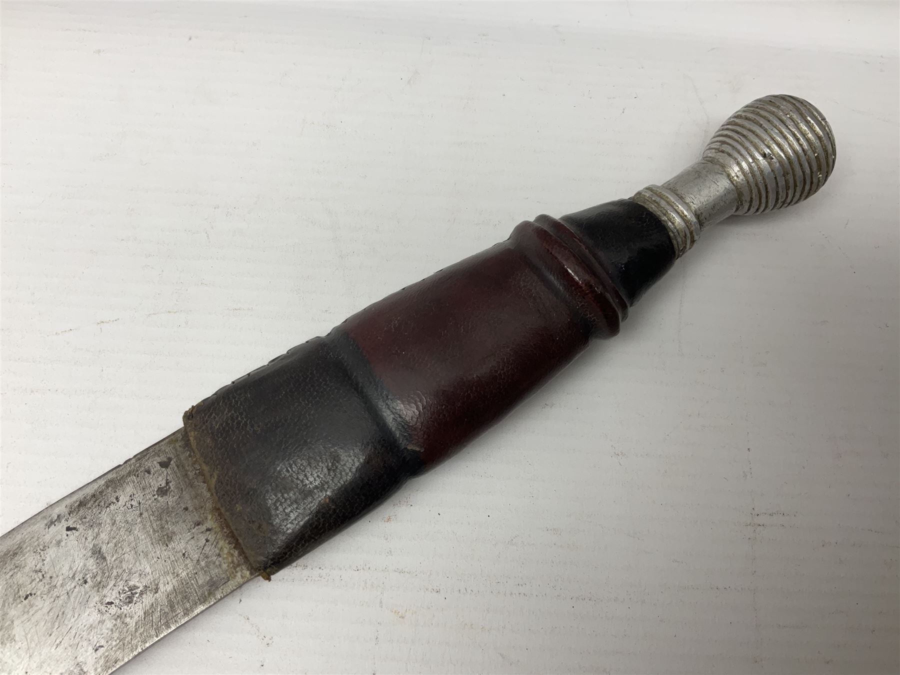 Mandingo sword with 63cm plain curving steel blade and leather covered grip with white metal ribbed spherical pommel; in decorative leather leaf scabbard with plaited tassels and shoulder strap L78cm overall