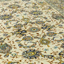 Central Persian Kashan hand-knotted carpet, ivory ground, with an all-over floral design of palmettes, scrolling vines and flowerheads in blue, ochre and beige tones, enclosed by a sage green border with repeating floral sprays and leafy scrolls, flanked by narrow guard stripes, with cotton fringes