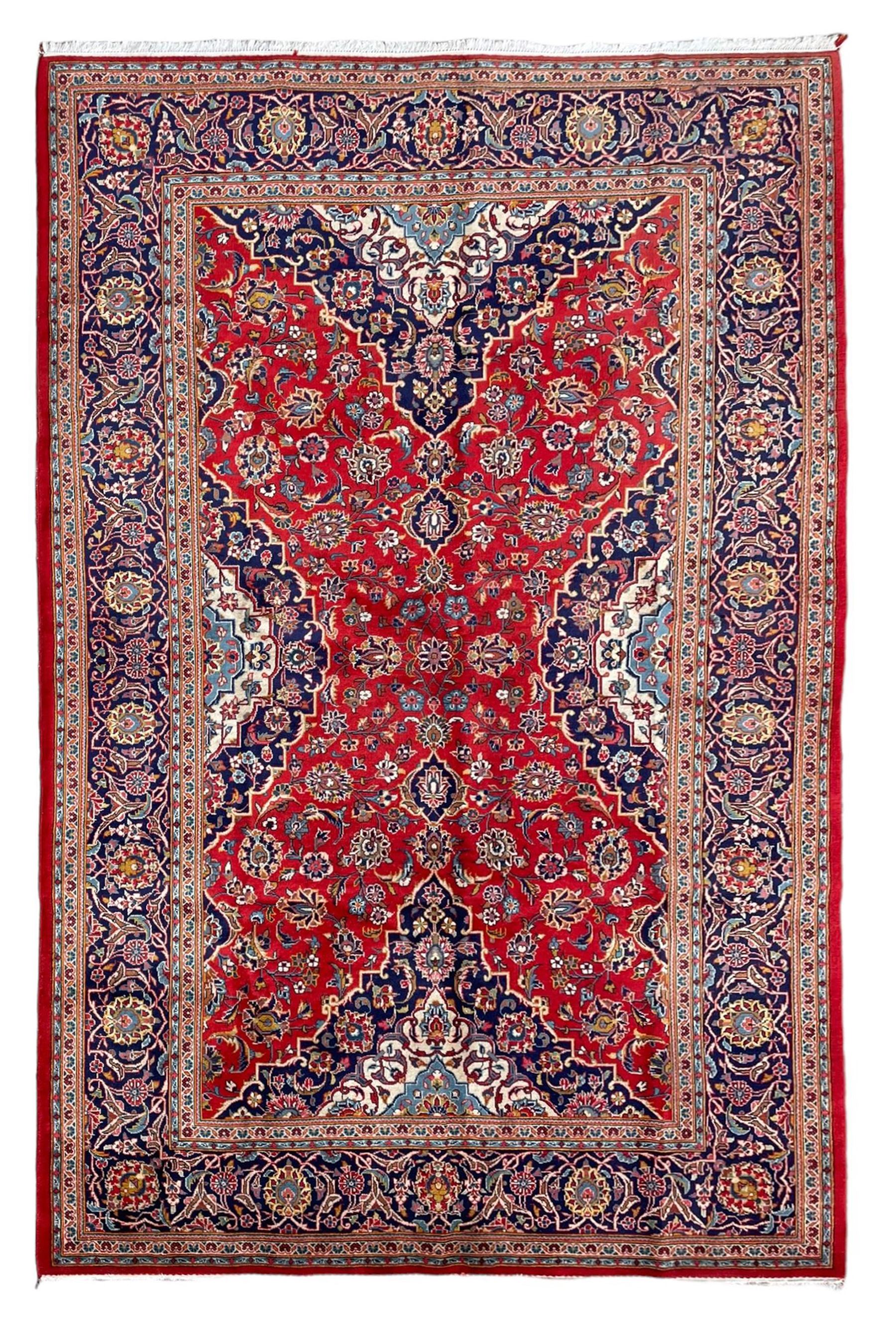 Persian Kashan crimson ground rug, overall arabesque design, the field decorated with swirled leafy branches and palmettes, indigo ground border decorated with repeating pattern, within guard stripes 