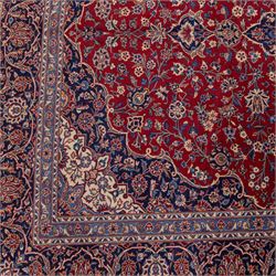 Central Persian Kashan hand-knotted carpet, madder red field with central cusped medallion in navy and ivory enclosing a floral rosette and radiating scrolling arabesques and palmettes, across a densely ornamented ground of finely drawn vinery enriched with a profusion of flowering sprays and stylized blossoms, spandrels in deep indigo and principal border of navy ground decorated with a continuous palmette and vine-scroll flanked by multiple guard stripes, with cotton fringe ends