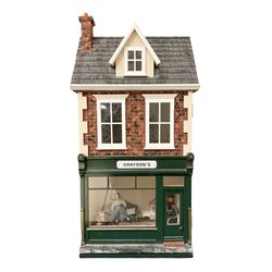 Good quality late 20th century dolls house by M. James dated 1996 in the form of 'Graysons' Shop with two storeys above; textured brick walls under a faux slated pitched roof with dormer window, hinged along the ridge giving access to a single attic room; the hinged front elevation with large glazed shop window and opening customer door; single room shop with first floor room over currently furnished with various scale domestic items including settee and armchair, longcase clock, chests of drawers, tables, chairs etc; together with bakery stock of cakes, pies, bread loaves and biscuit tins W39cm H79.5cm D33cm