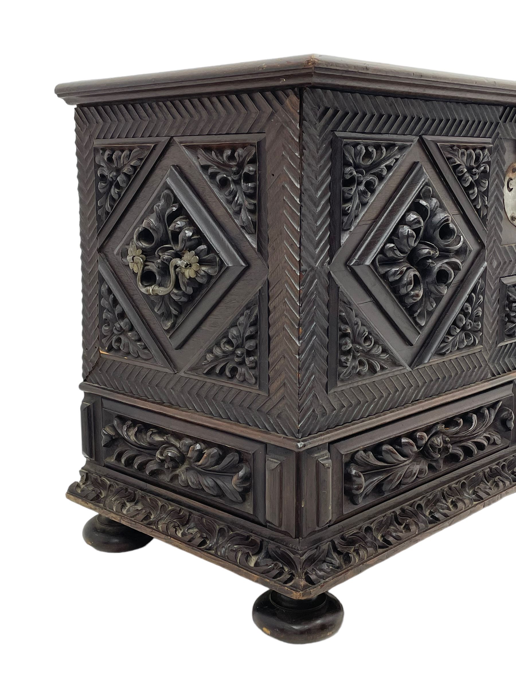 Early 20th century carved hardwood kist or mule chest of small proportions, moulded rectangular hinged lid over foliage carved geometric lozenge panels, the frame carved with chevron banding, fitted with two drawers, the drawer fronts carved with extending leaf motifs over a foliage carved lower moulding, on turned feet