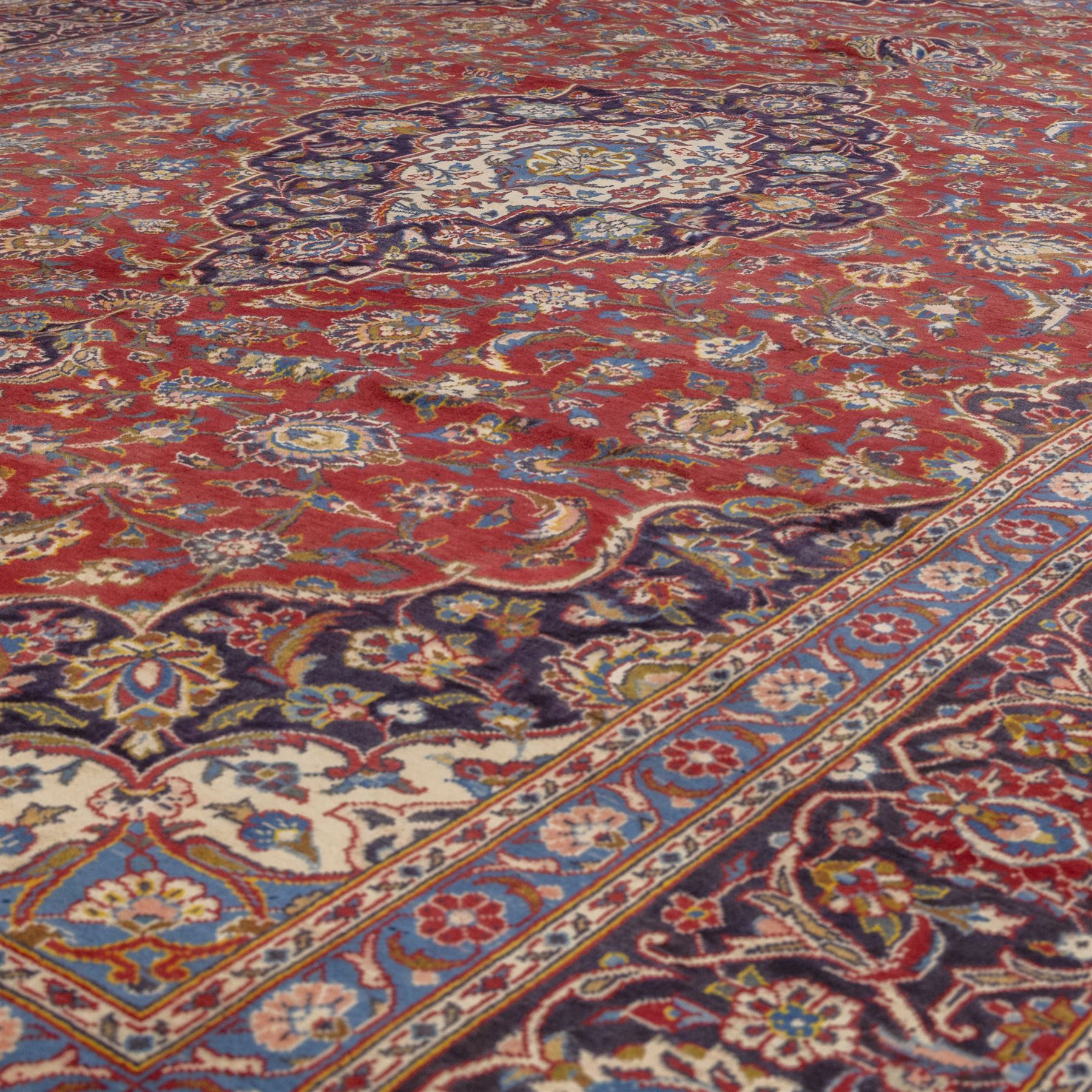 Central Persian Kashan hand-knotted carpet, madder red field with a central cusped medallion of ivory and deep indigo ground, enclosing a stylized floral rosette, framed by scrolling arabesques, field richly ornamented with an intricate network of flowering vinery, palmettes, and delicate floral sprays, spandrels in deep indigo, principal border of navy ground decorated with a continuous palmette and vine-scroll, flanked by multiple guard stripes, with cotton fringe ends 