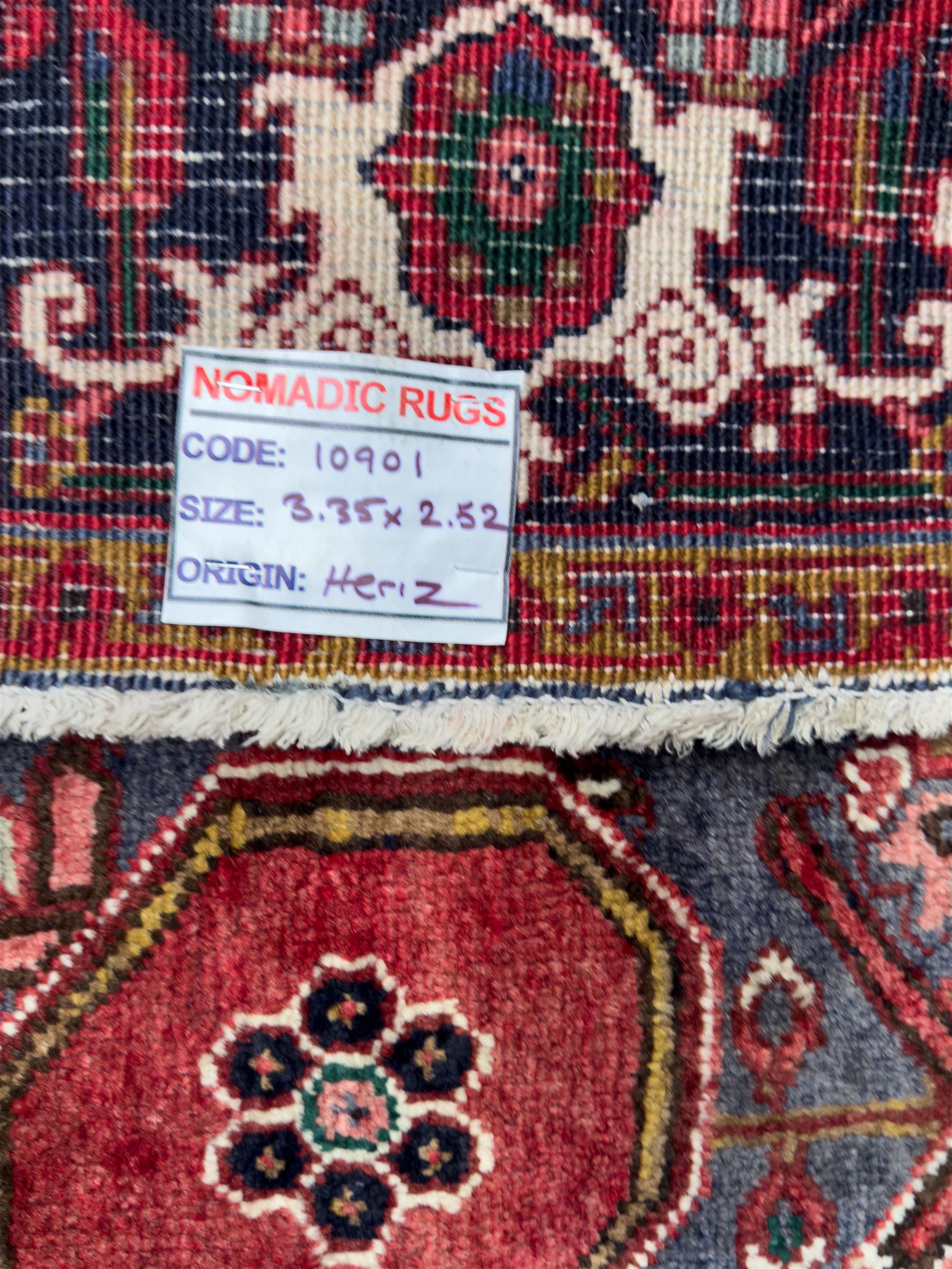Northwestern Persian Heriz hand-knotted carpet, madder red field with a bold stepped medallion in indigo, ivory, and ivory, surrounded by angular floral motifs and stylized leaves, and the spandrels set within a dark border of rosettes and angular vine motifs, 335cmx252cm