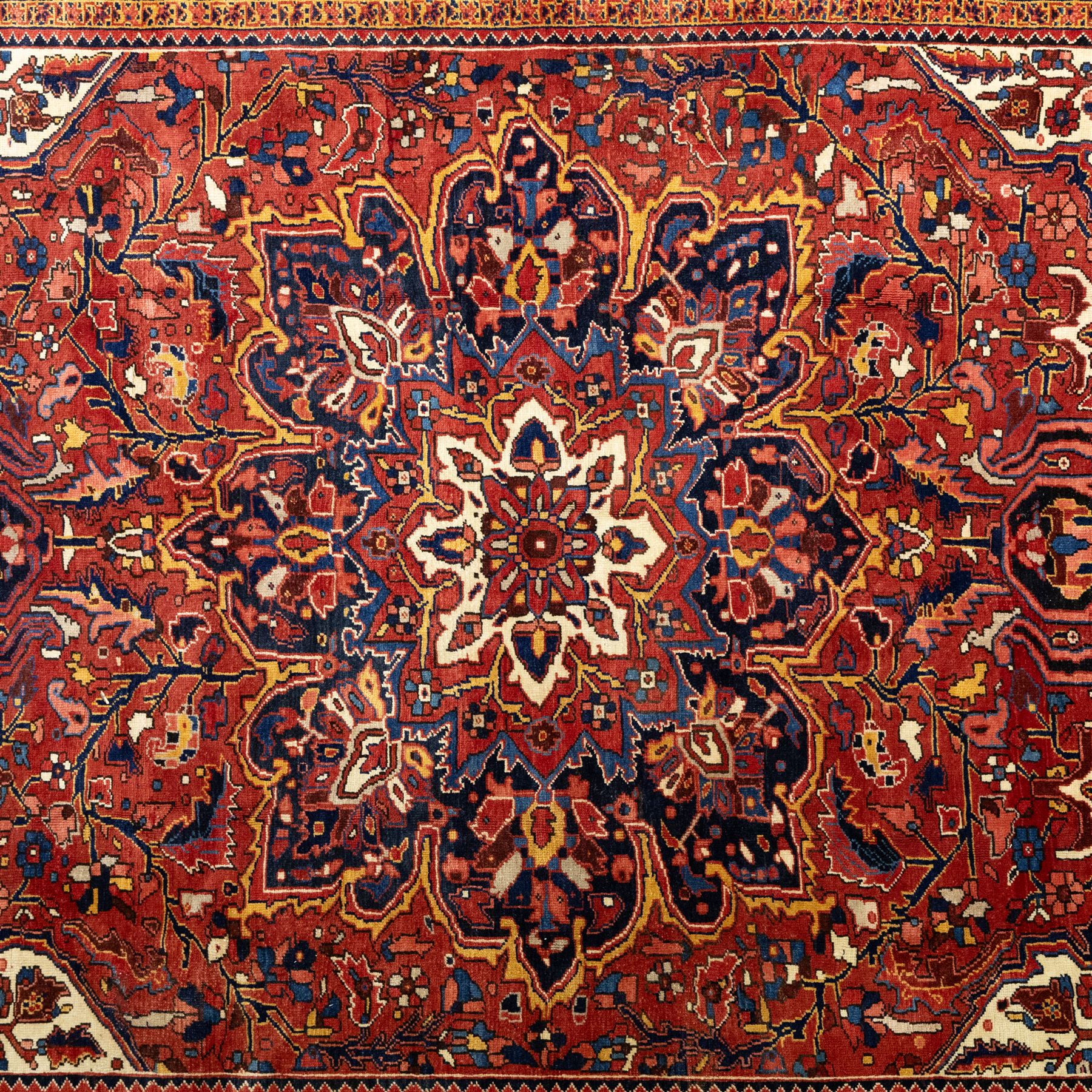 North West Persian Ahar hand-knotted carpet, red field with large central angular star medallion with stepped outlines and radiating geometric motifs surrounded by stylized floral and serrated leaf forms, enclosed by a deep blue main border decorated with angular palmettes and rosettes flanked by multiple guard stripes, with cotton fringe ends 