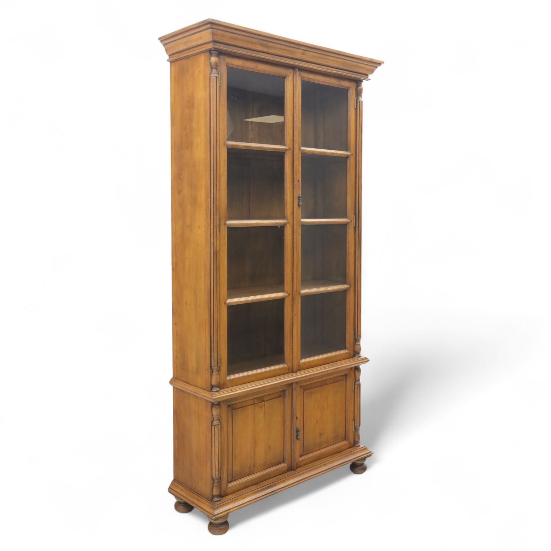 Pair of late 20th century Thai hardwood display cabinets, each with moulded cornice over twin glazed doors enclosing adjustable shelves, panelled cupboard doors below flanked by fluted columns, raised on bun feet