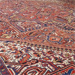 North West Persian Heriz hand-knotted carpet, madder red field, with a large angular stepped central medallion with serrated outlines, flanked by bold stylized palmettes, hooked leaves and geometric devices, spandrels similarly decorated within a wide geometric main border featuring angular floral motifs, enclosed by multiple guard borders in madder red, indigo blue, ivory, olive green and soft ochre, with cotton fringe ends 