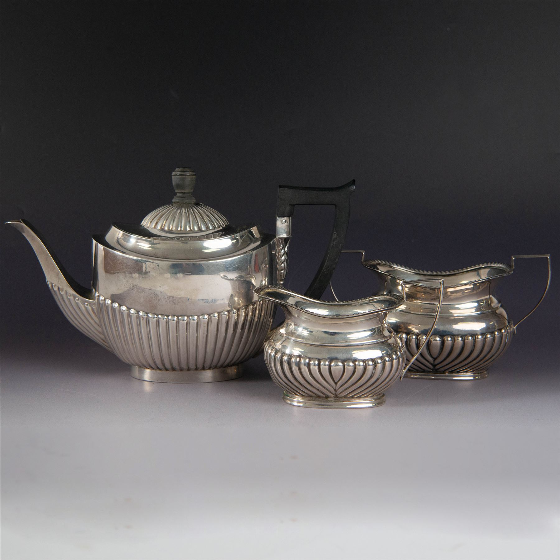 Victorian silver bachelors teapot, of part fluted oval form with ebonised wooden handle and finial, hallmarked Thomas Bradbury & Sons, London 1896, together with a Edwardian silver milk jug and open sucrier, of similar form with oblique gadrooned rims, hallmarked George Unite, Birmingham 1902 & 1906