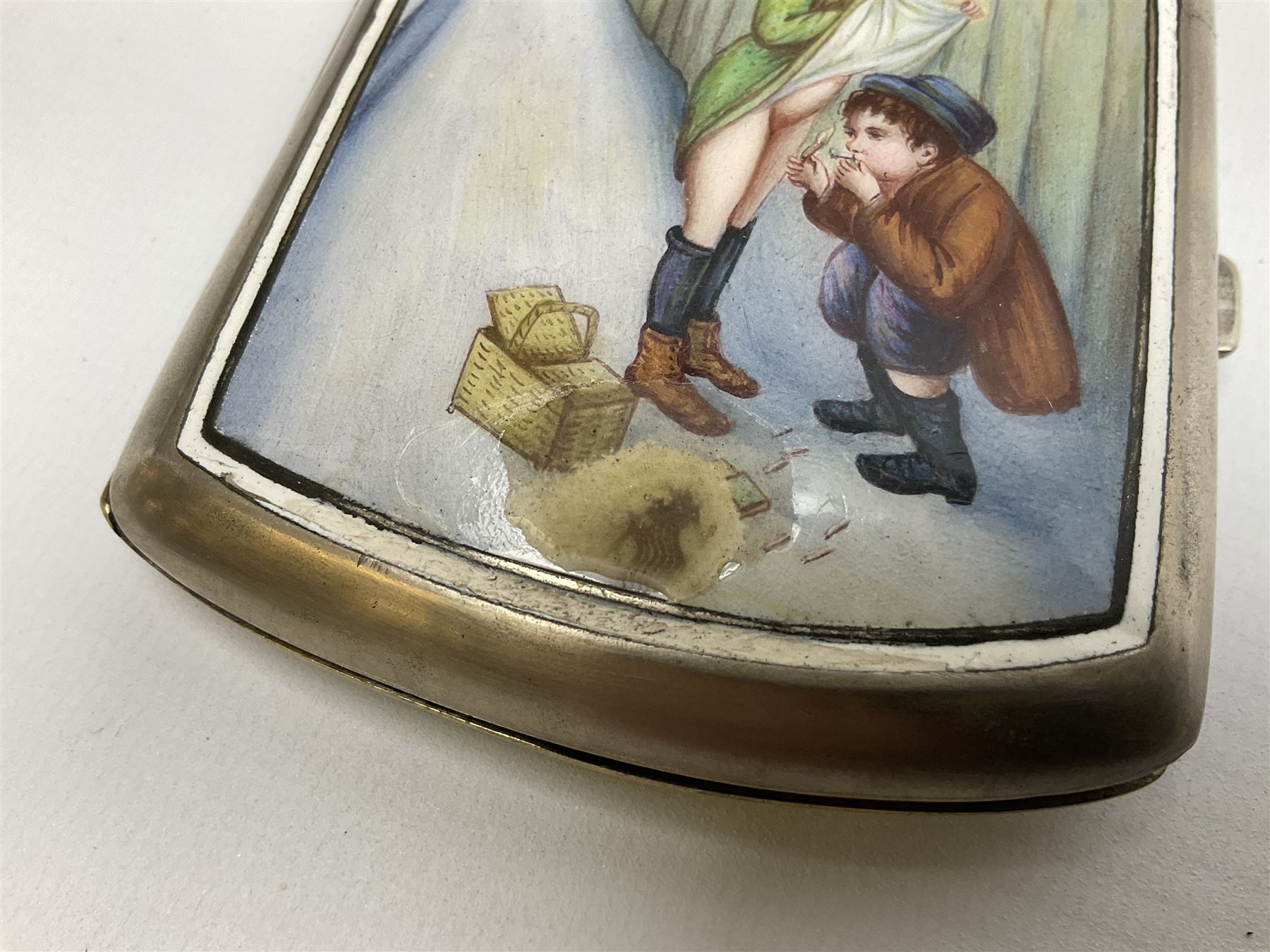 Early 20th century Continental erotic cigarette case, the silver-plate body of curved rectangular form with cover decorated with painted enamel street scene depicting young boy in squatting position lighting a cigarette shielded from the wind by his girlfriend, who is lifting the skirt of her dress up over him, with gilt interior, L9cm