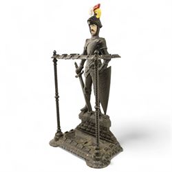 Early 20th century painted cast iron hall stand, formed as a standing armoured figure with red and yellow feathered crest, twin rails and integral drip tray with castellated edge