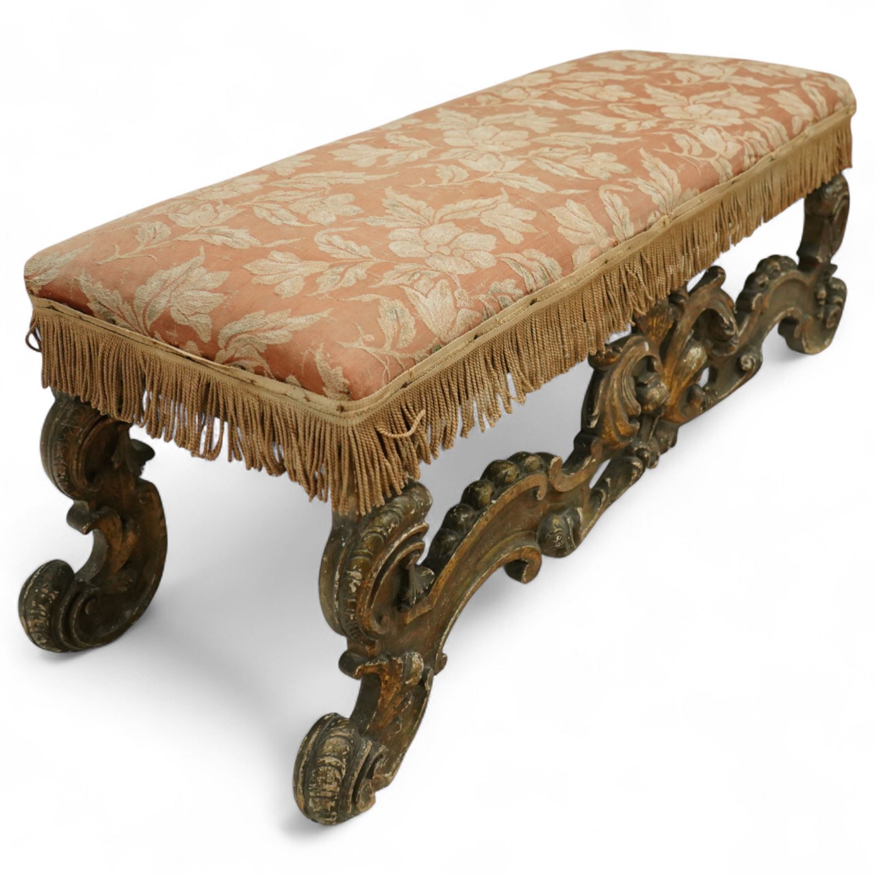 19th-century Rococo-style giltwood window seat, upholstered in floral-patterned coral-ground fabric with fringed border, the intricate scrolled supports united by pierced scrolling foliate stretchers with acanthus and C-scroll decoration