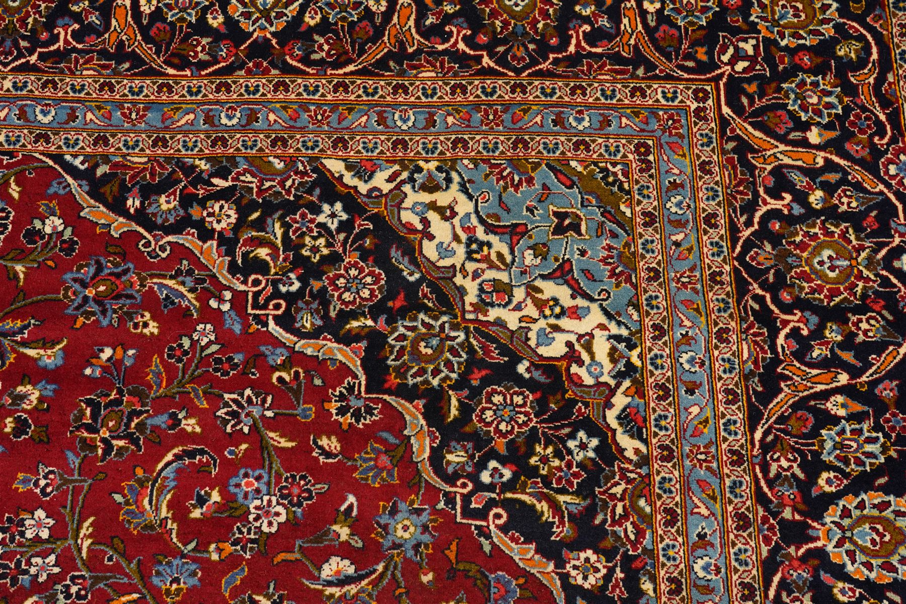 Central Persian Kashan hand-knotted carpet, crimson ground field, with central cusped lozenge medallion, enclosing a palmette and a floral rosette arrangement, surrounding field densely populated with scrolling arabesques, flowering vinery and stylized boteh motifs in tones of indigo, pale blue, ivory and ochre and spandrels of confirming design, flanked by multiple guard stripes, with cotton fringe ends 