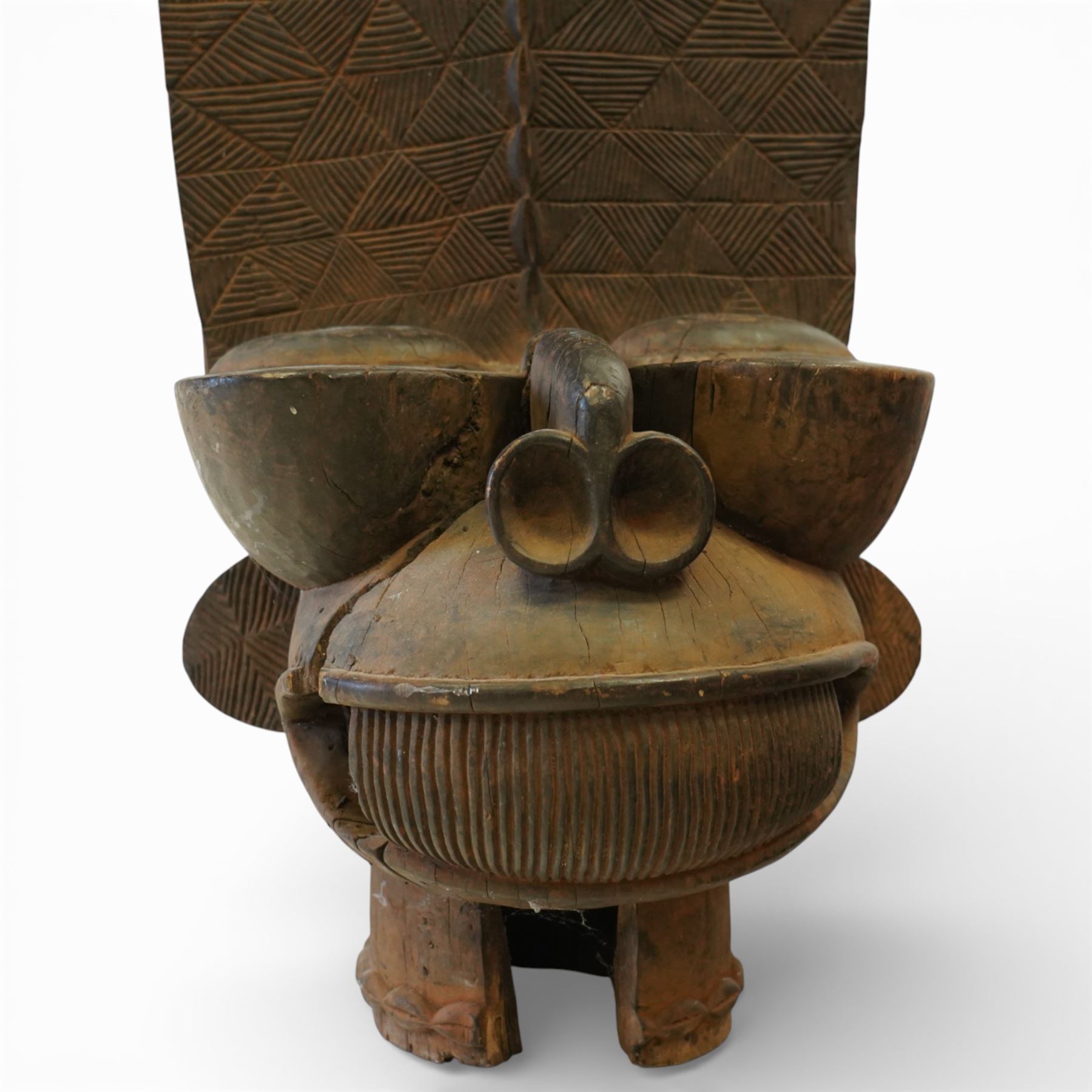 African carved hardwood ceremonial seat, monoxylous construction with twin dished seat wells, the tall back incised with repeating geometric triangular motifs, the front carved in the form of a tribal mask with projecting features, raised on a cylindrical base with ribbed and textured carving