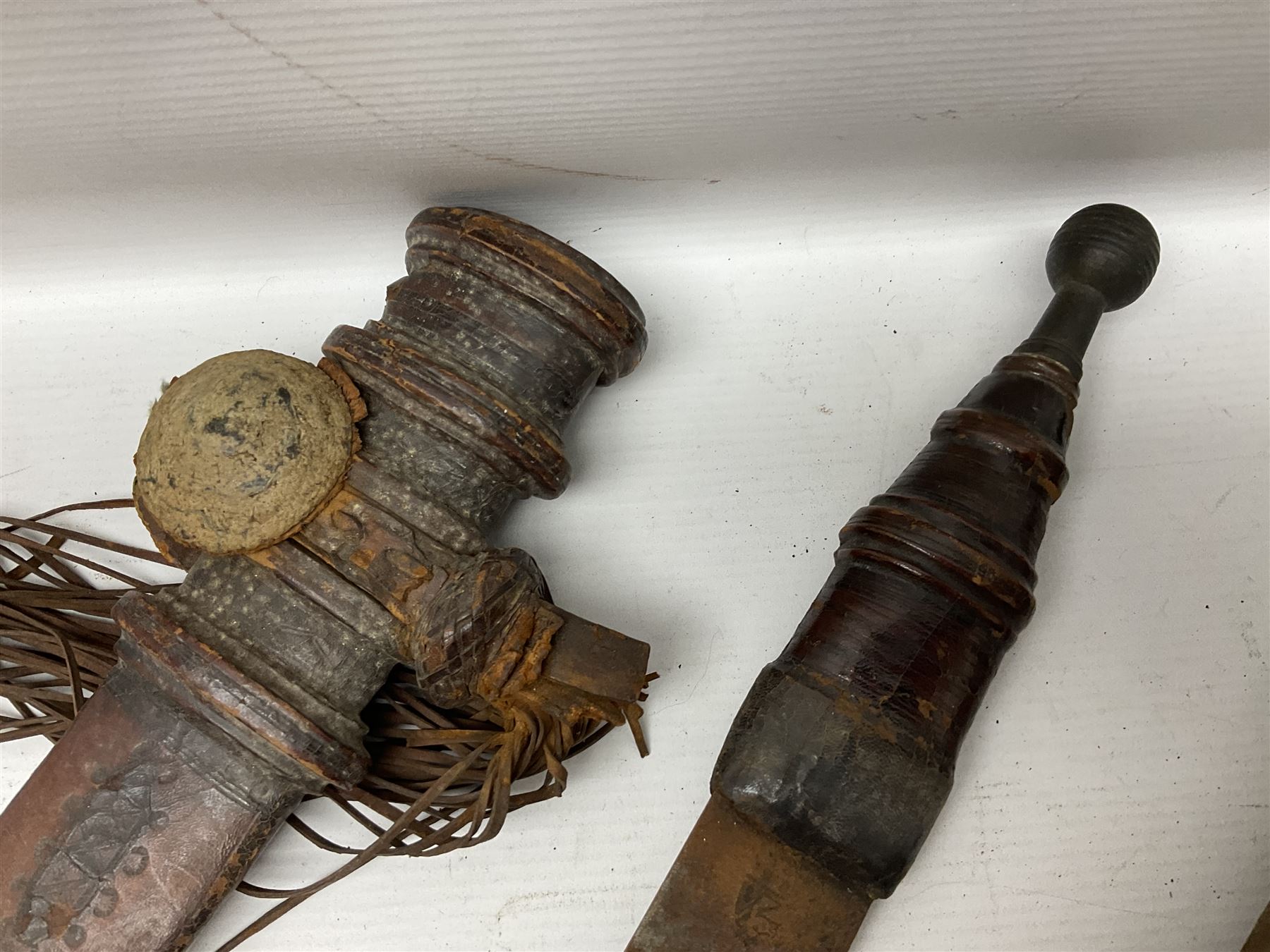 Mandingo sword, the 63cm plain curving steel blade marked with a horses head and the initials G.S.; leather covered grip with metal ribbed spherical pommel; in decorative leather leaf scabbard mounted with tassels and roundels L84cm overall; and a North African flyssa sword in poor condition with wooden scabbard (2)