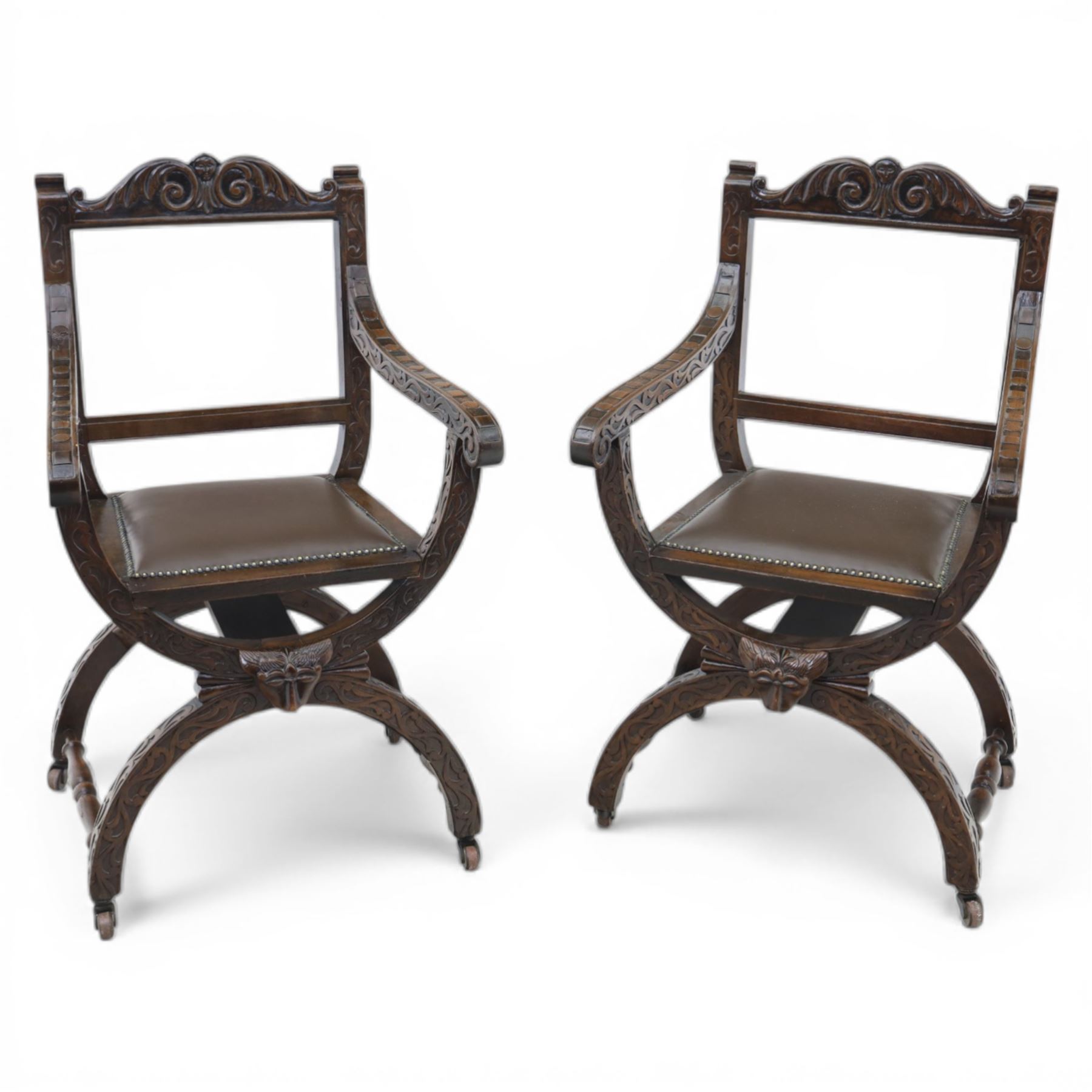 Late 20th century stained beech Savonarola chair, the carved crest rail and open arms above an upholstered leather seat, raised on an X framed base with carved foliate and mask decoration, joined by turned stretcher and ending in castors