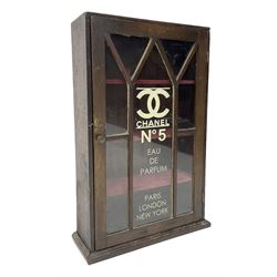 Early 20th century wall hanging cabinet with glazed panel door, decorated with later cream Chanel No.5 advertising, the hinged door opening to reveal three shelves upholstered with berry red fabric, H51.5cm