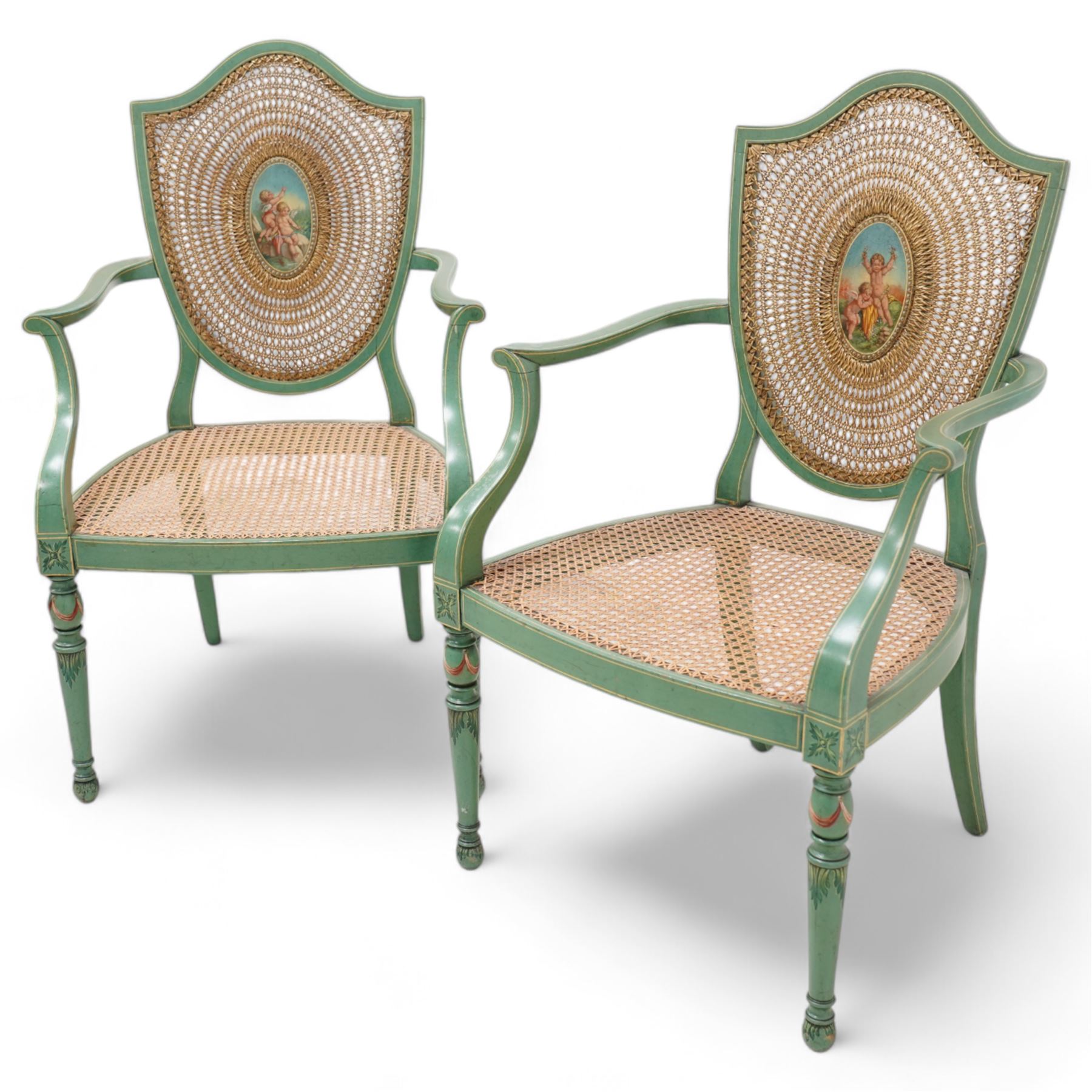 Pair of Edwardian Hepplewhite Revival green-painted open armchairs, cane work shield back with central oval panel panted with putto set upon rocky landscape with water and trees, shaped and rolled arms enclosing cane work seat, on turned front supports decorated with acanthus leaves and draped festoons 