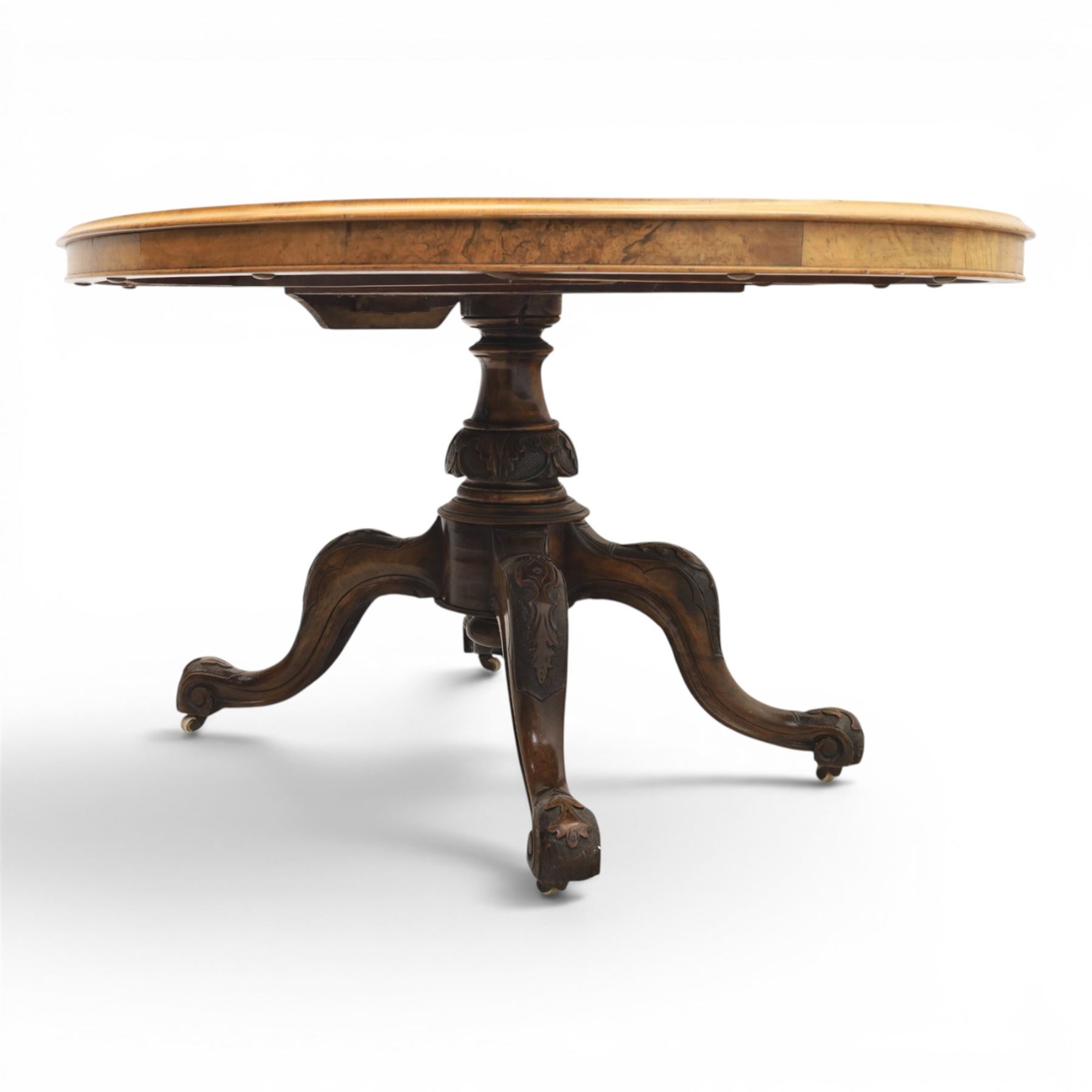 Victorian figured walnut oval loo tilt top table, raised on a heavily carved quatriform base with scrolled supports, terminating in castors