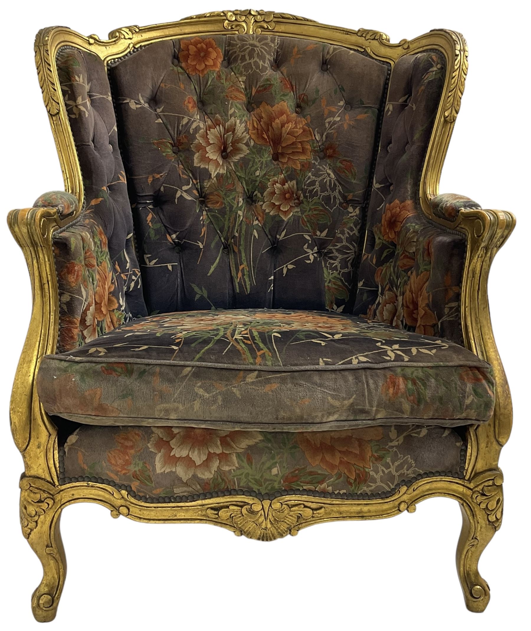 Pair of Louis XVI design gilt framed wingback armchairs, cresting rail carved and moulded with foliate decoration and C-scrolls, upholstered in buttoned floral patterned mauve velvet with loose seat cushion, scallop carved apron over cabriole supports