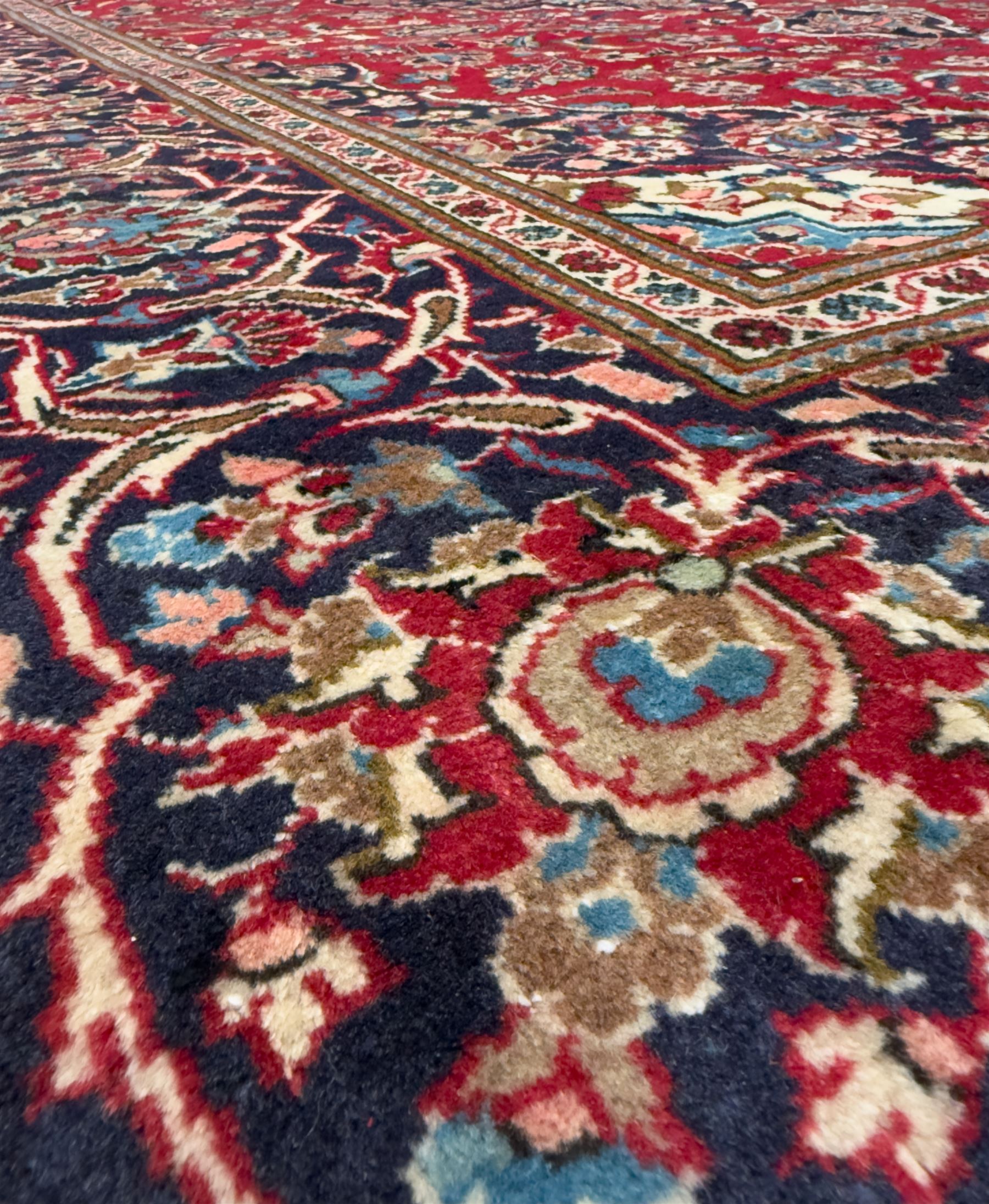 Central Persian Kashan hand-knotted carpet, the deep red ground with a central lozenge medallion in ivory and navy, surrounded by dense scrolling floral and vine motifs, navy spandrels and a wide border with stylised palmettes and flowerheads enclosed by multiple guard stripes