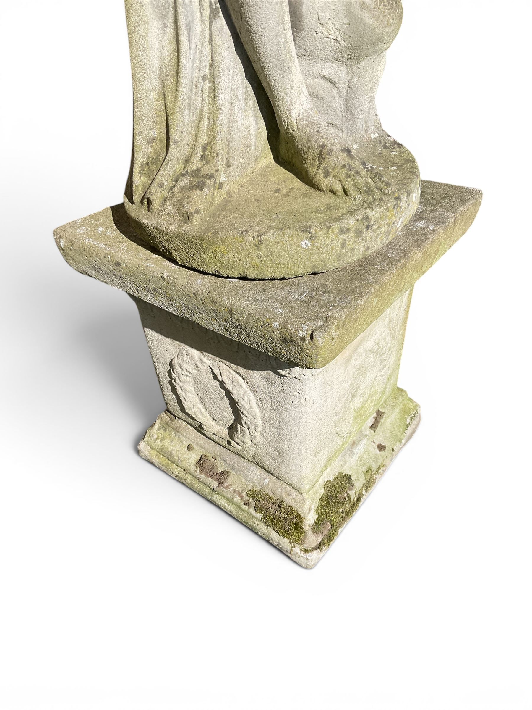 Cast stone garden figure of a classically draped nude female inspired by Aphrodite statues, with stylised up-do arranging her chitin on a circular base, the rectangular pedestal with egg-and-dart moulding over a floral wreath, in weathered finish