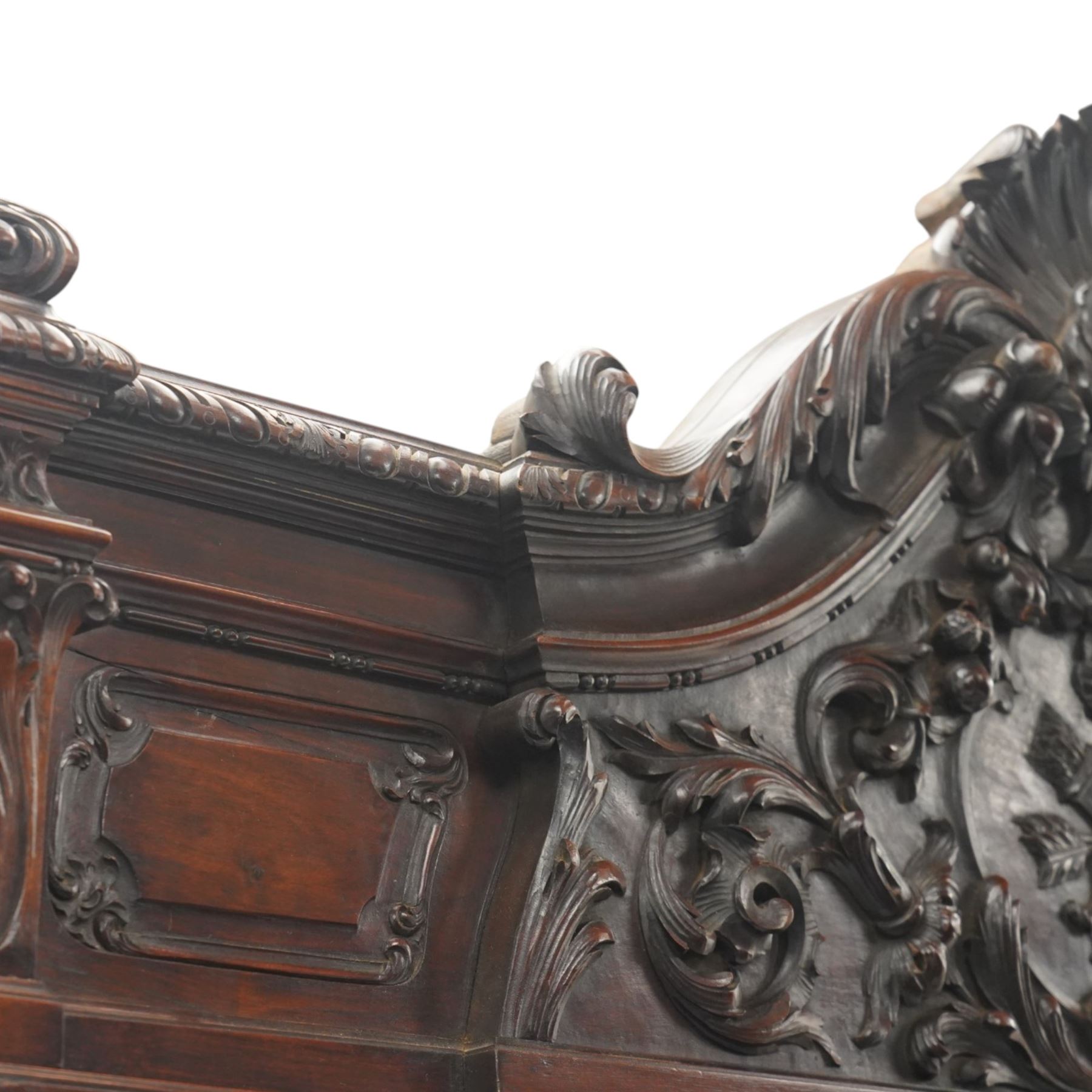 Large and impressive 19th century heavily carved mahogany display cabinet, the ornate pediment carved with a central scalloped and foliate rocaille motif with extending acanthus leaves and egg-and-dart moulding flanked by acanthi finials, over a central cartouche with carved bow-and-arrow motif and trailing scrollwork, the bookcase enclosed by two glazed doors with bevelled glass sides, the base with a gadrooned upper edge over a central rocaille ornament with scrolling acanthus leaves, the panelled frieze with further foliate motifs, raised on large carved snarling lion mask supports with floral garlands and claw feet, united by a concave rectangular platform base with gadrooning egg-and-dart moulding