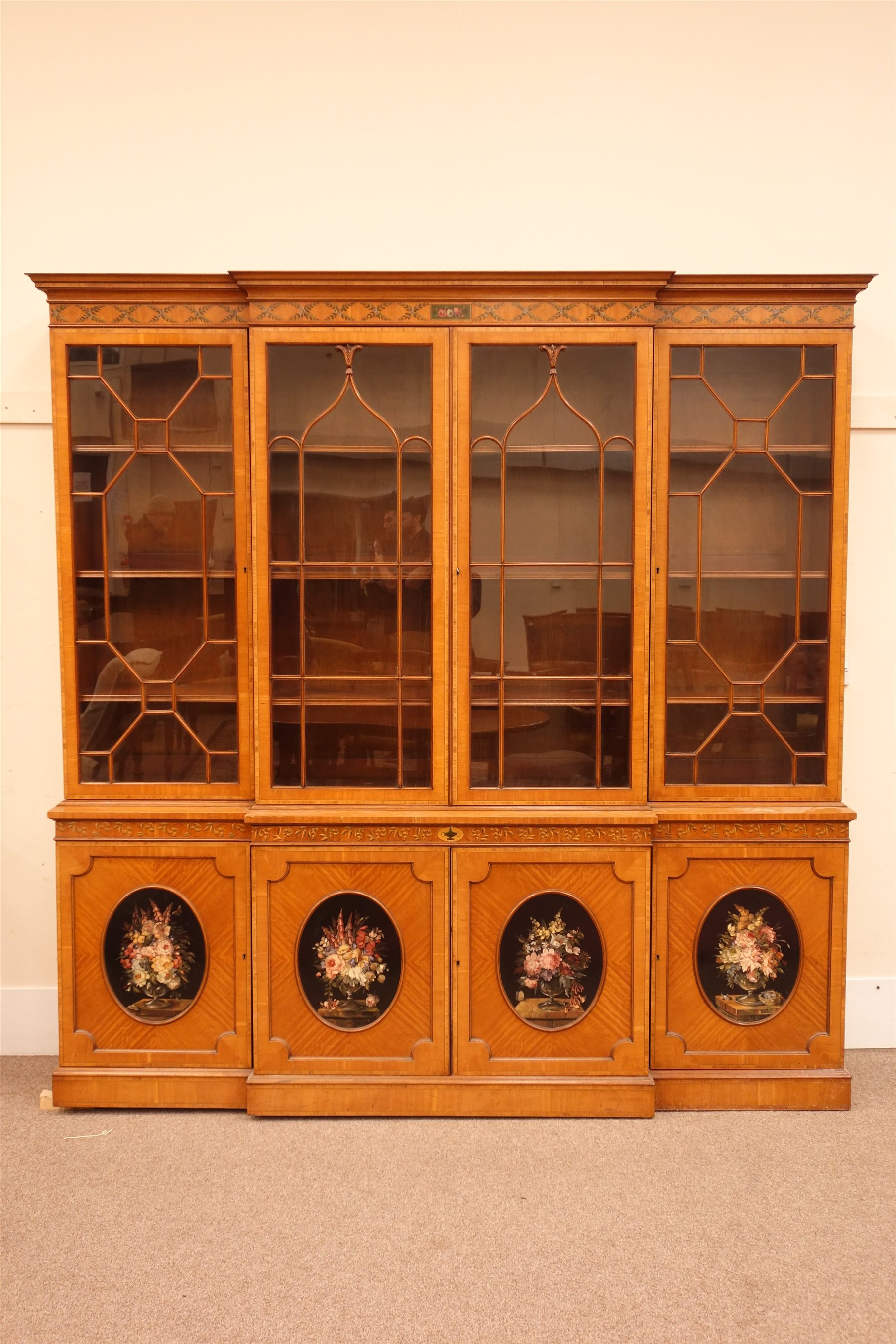 Sheraton Revival satinwood breakfront bookcase, frieze painted with scrolling leafage above four astragal doors with adjustable shelves, the base with four doors painted with floral sprays in oval panels, on a plinth base, W234cm, H235cm, D52cm