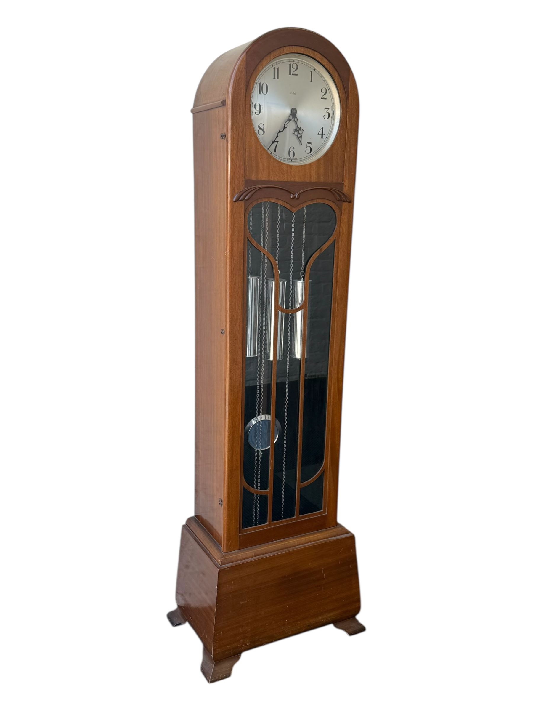 English- Art deco mahogany 8-day longcase clock, with an arched top and fill length fully glazed door beneath, on a short plinth with bracket feet, three train chain driven movement with visible stainless steel cased weights and pendulum, silvered dial with Arabic numerals, minute track and pierced steel hands, quarter chiming movement with  Westminster/ Whittington/ strike/silent selection.
