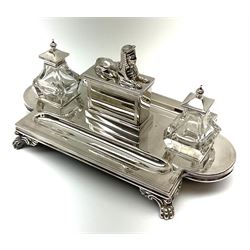 A 20th century silver plated desk stand, the shaped base with twin pen trays, upon four paw feet, supporting a central wafer box with sphinx surmount to the cover and engraved dedication to front, flanked by twin glass inkwells with hinged silver plated covers, L28cm 