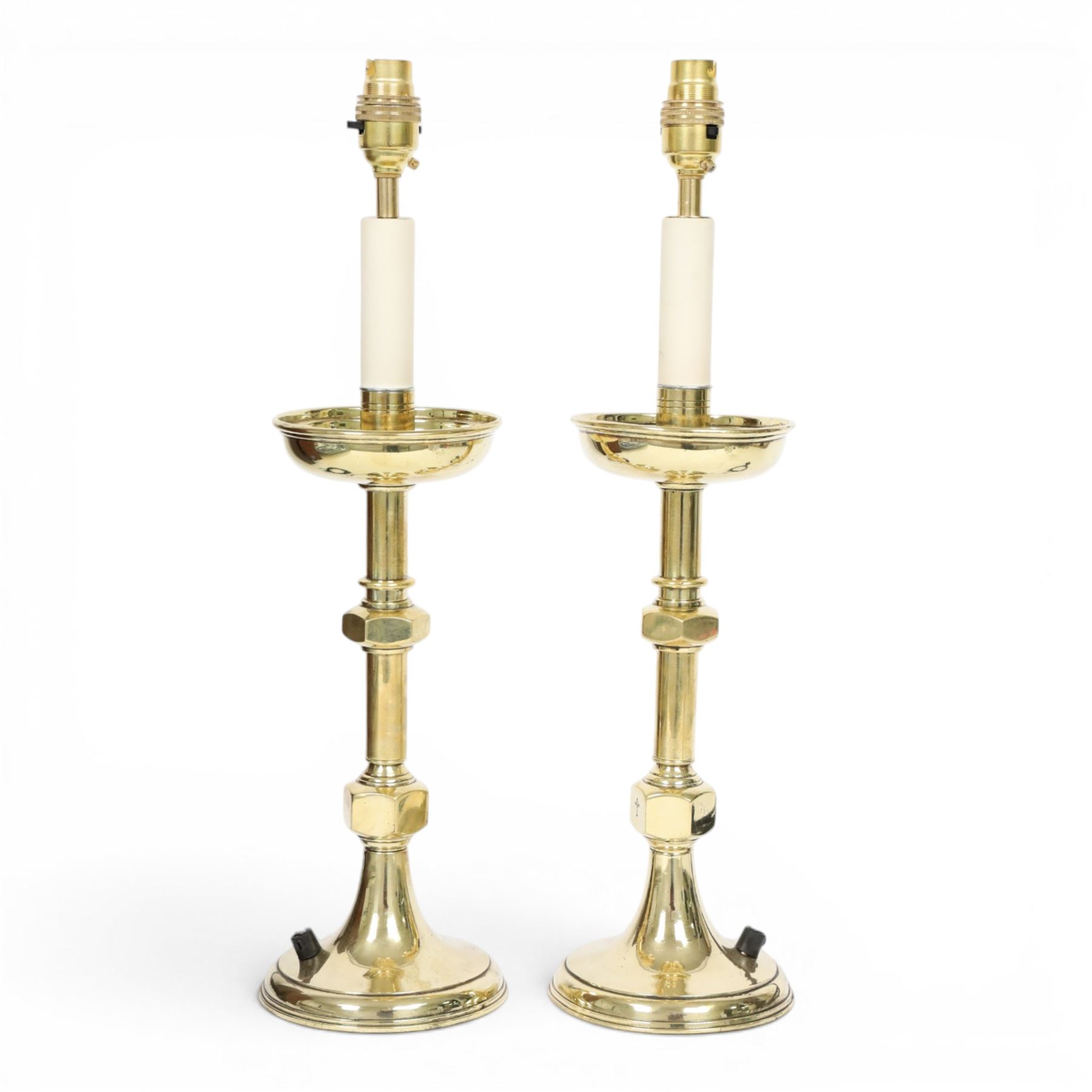 Pair of early 20th century brass alter candlesticks converted to table lamps, each with stepped and spreading circular base leading to a double hexagonal knopped column, and plain socket above inverted domed circular drip pan, the socket with light fitting, overall H49cm