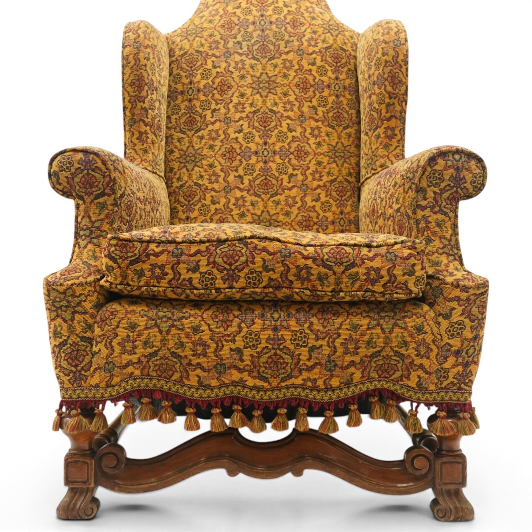 Pair of William & Mary design wingback armchairs, the arched high-back over rolled arms with a loose seat cushion and sprung seat, upholstered in old-gold ground foliate patterned fabric, raise on scroll feet united by a front serpentine stretcher