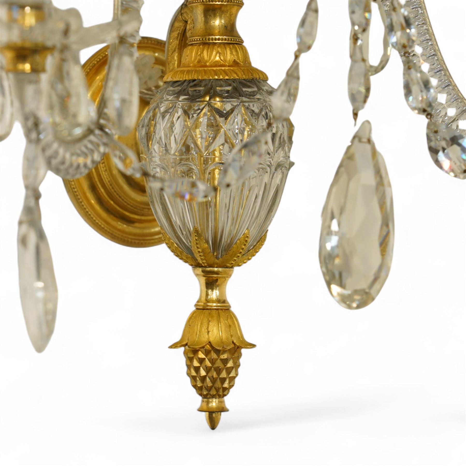 Pair of George III ormolu and cut-glass wall-lights, each with a tall central urn-shaped finial above a series of faceted glass spires and scrolled branches, the ormolu-mounted nozzles and drip pans hung with festoons of cut-glass drops and pear-shaped pendants, the lower section centred by a pineapple finial, wired for electricity