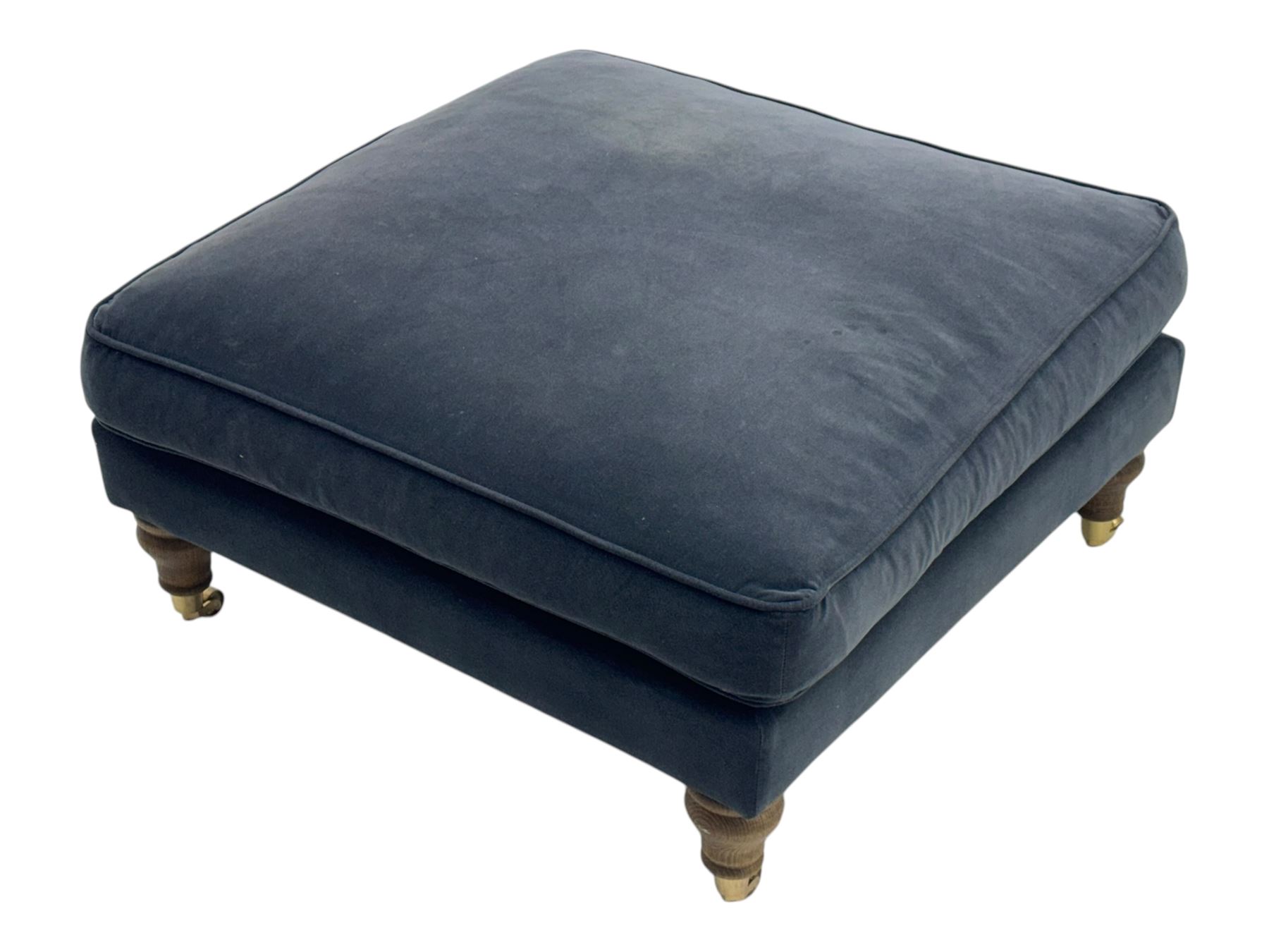 Contemporary square footstool upholstered in navy armour smart velvet, featuring a deep padded top, turned wood supports, and brass castors